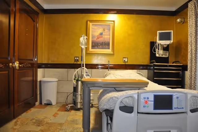 A medical patient room with a hospital bed, an IV stand, a bedside table, medical equipment mounted on the wall, a trash bin, and a framed picture of an American flag on a yellow wall.
