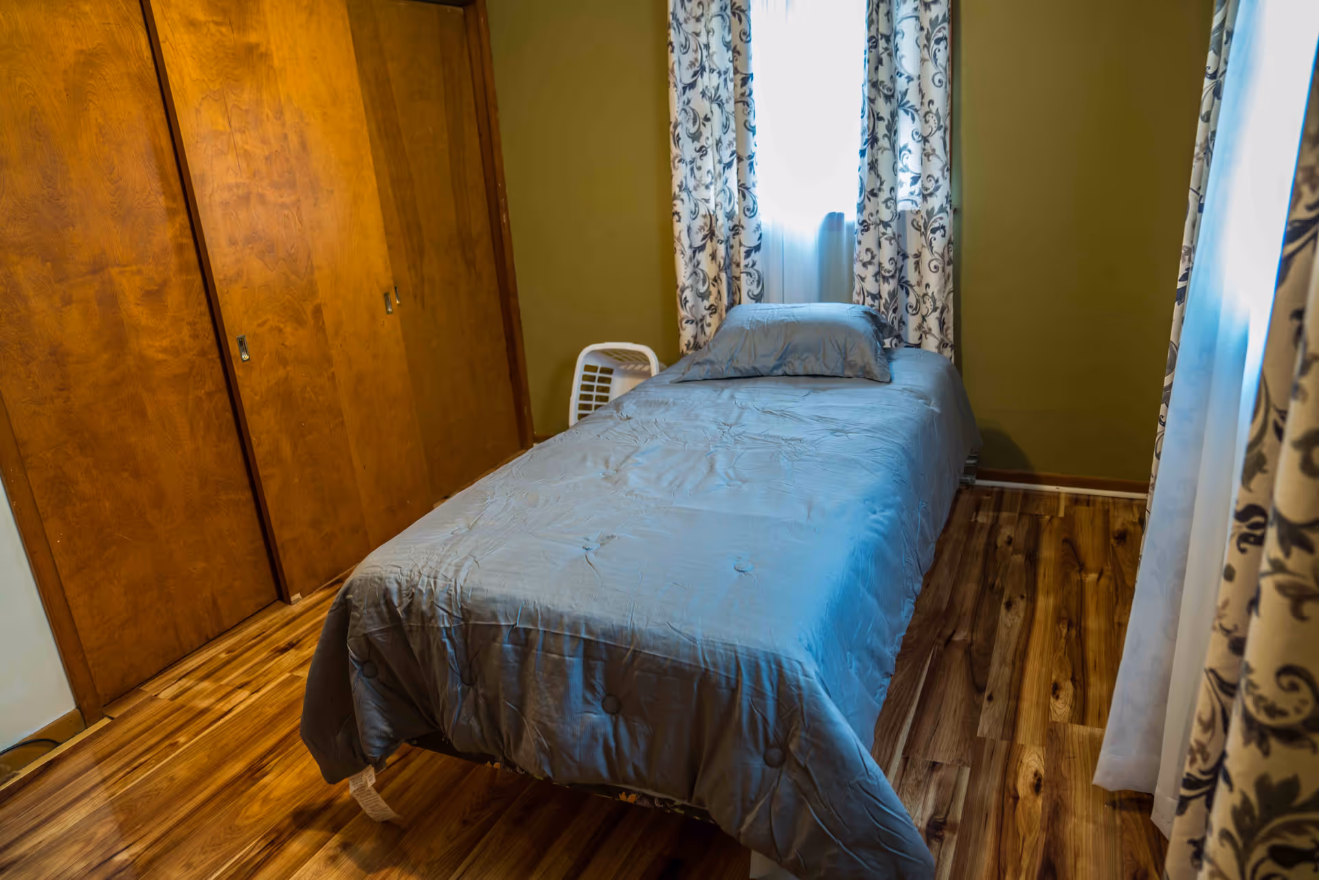 A small bedroom with a single bed covered in a gray comforter and a matching pillow. The room has wooden floors, olive green walls, and a window with patterned curtains letting in natural light. There is a wooden closet with sliding doors on one side and a white laundry basket near the bed.