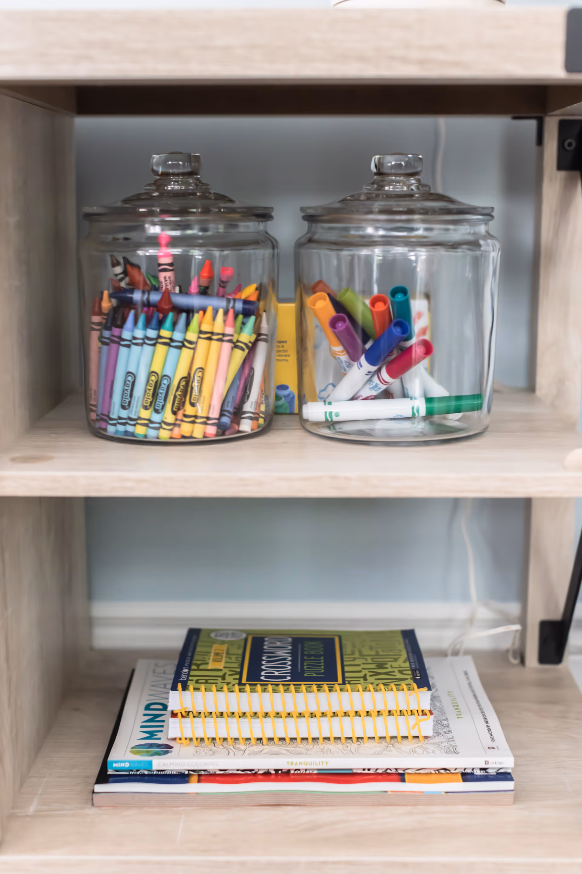 Glass jars filled with crayons and markers sit on a wooden shelf beside stacked notebooks.