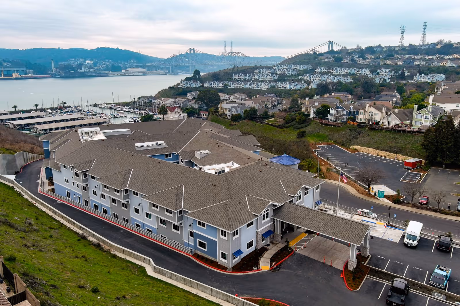 Aerial view of a large senior living facility building with a covered entrance and parking lot, situated near a waterfront with boats and a marina. Hills and a bridge are visible in the background under a cloudy sky.
