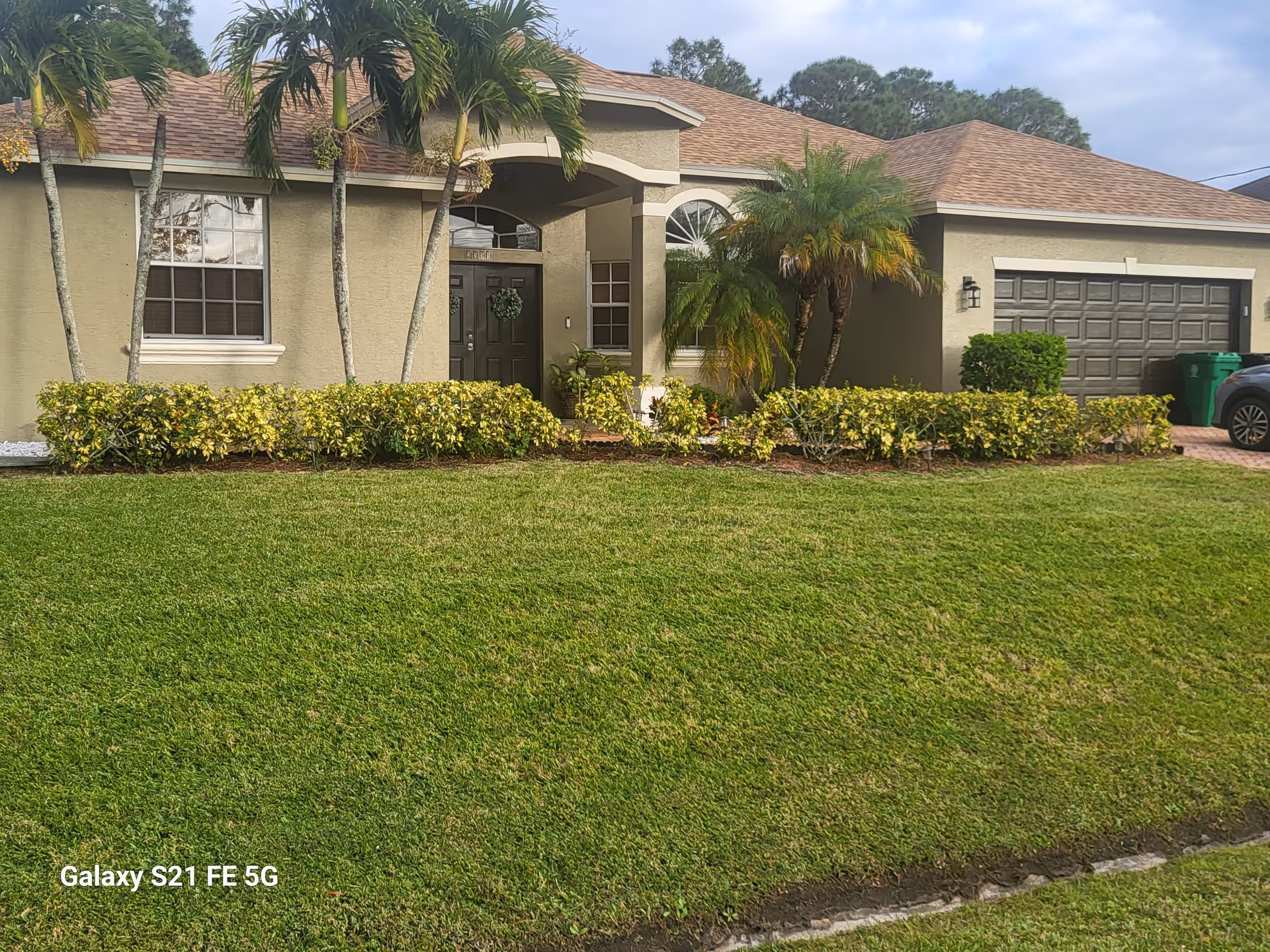 Front exterior view of a single-story beige house with a manicured lawn and palm trees.