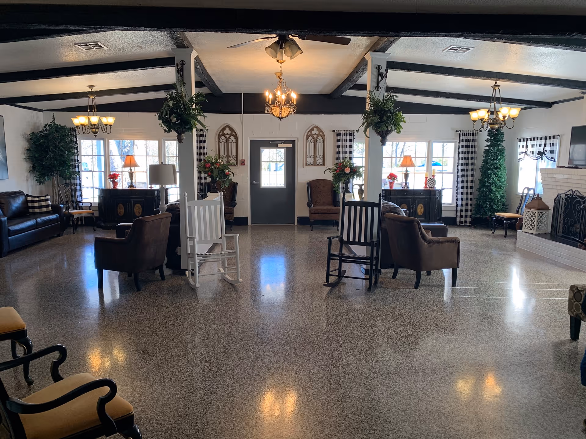 A spacious senior living facility common area with polished floors, multiple seating options including leather sofas, armchairs, and rocking chairs. The room features large windows with checkered curtains, decorative plants, chandeliers, and a white brick fireplace with a TV mounted above it. There are also side tables with lamps and floral arrangements, and ceiling beams adding to the cozy ambiance.