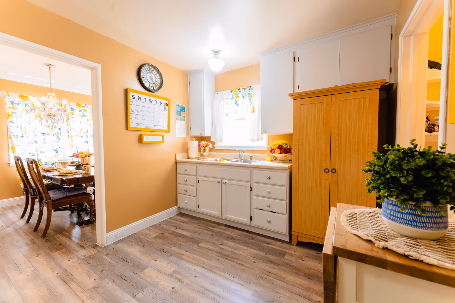A bright kitchen with white cabinets, a wooden floor, and a window with floral curtains above the sink. A wooden cabinet and a small table with a potted plant are on the right side. A wall clock and a weekly calendar are mounted on the peach-colored wall. Through an open doorway, a dining room with a wooden table, chairs, and a chandelier is visible.