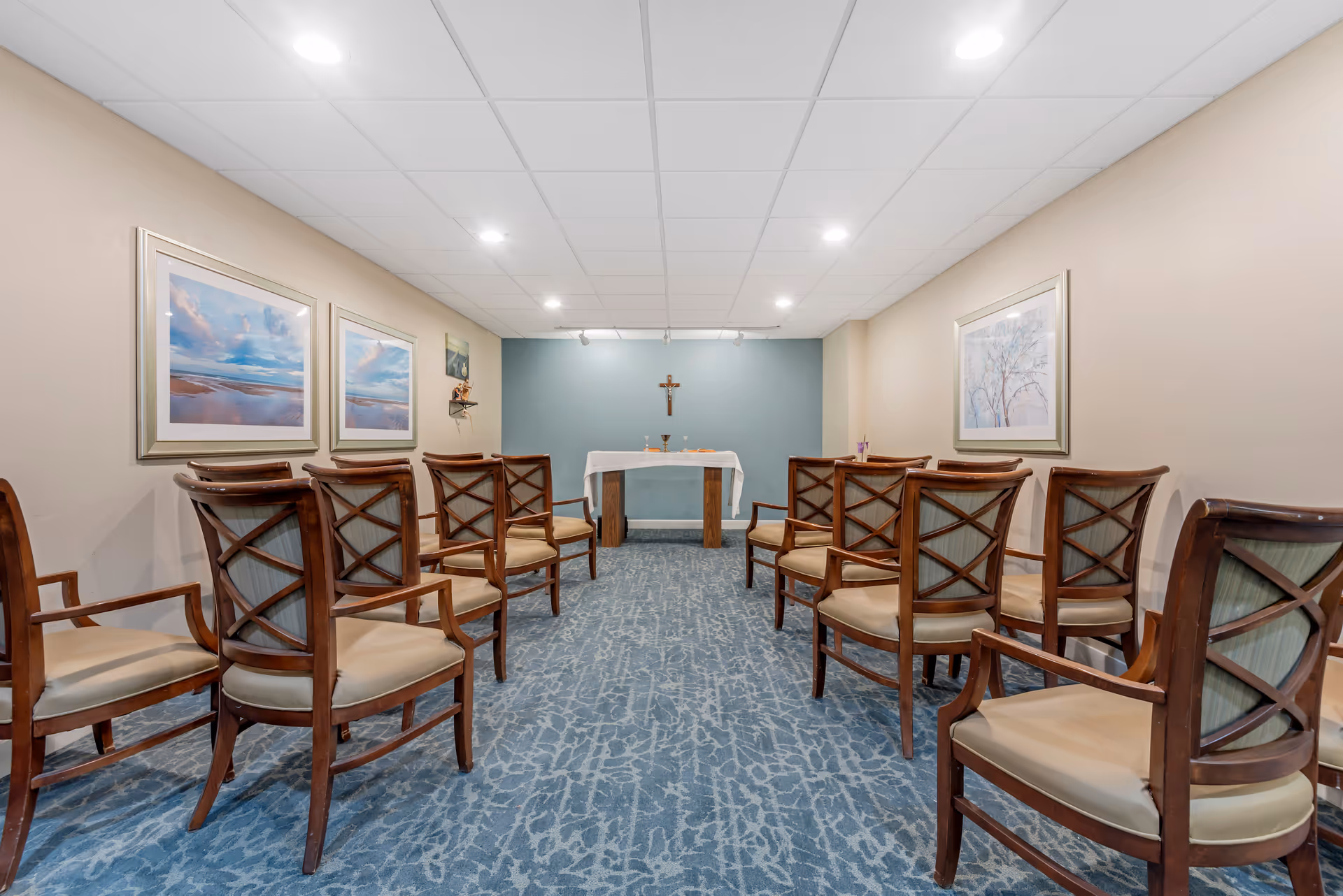 Small chapel-style meeting room with rows of wooden chairs facing a table altar and a cross at the front.