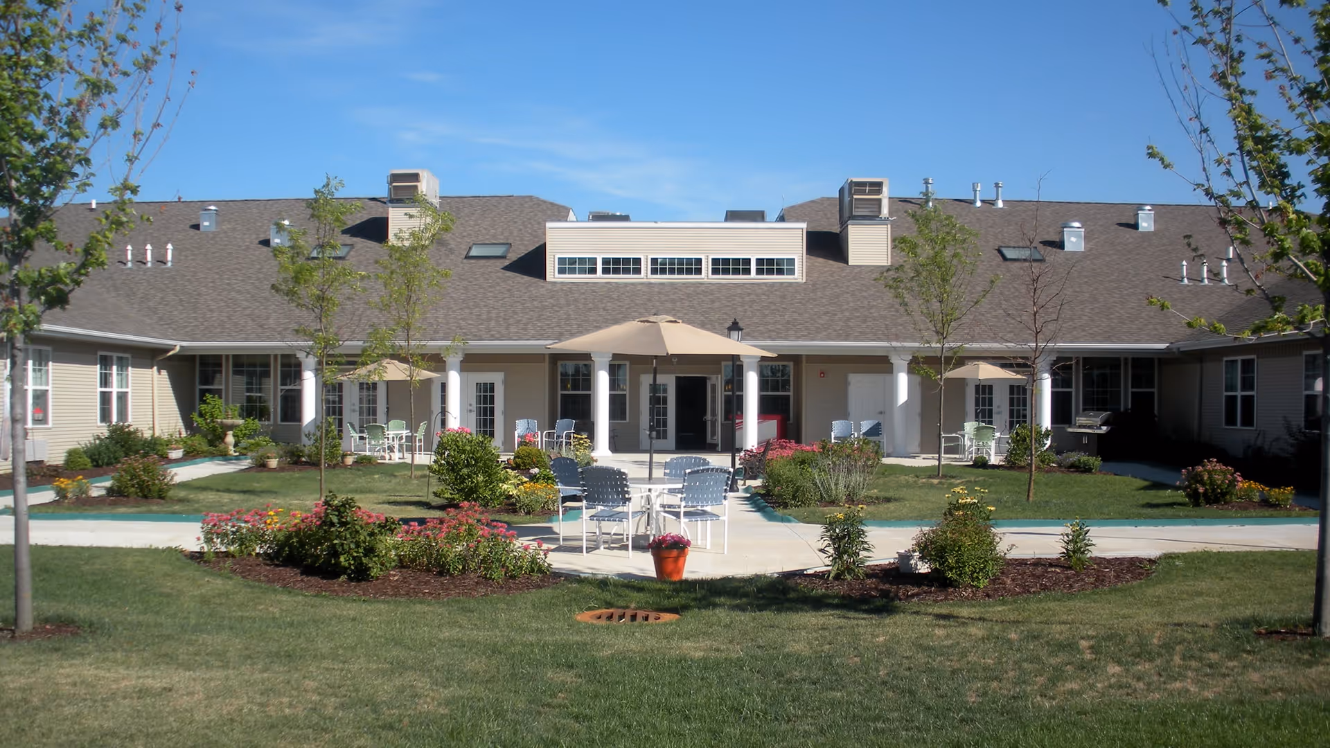 Courtyard patio with tables, umbrellas, and landscaped flower beds in front of a single-story assisted living building.
