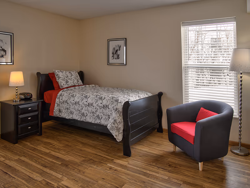 A furnished senior living bedroom with a dark bed, nightstand, chair, and large window.