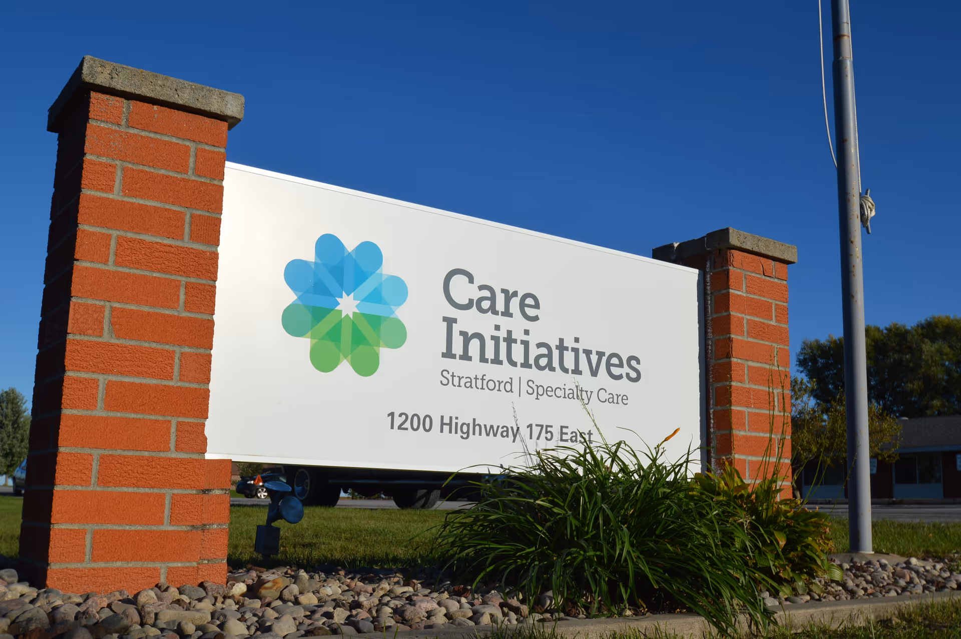 Outdoor sign for Care Initiatives Stratford Specialty Care located at 1200 Highway 175 East, mounted between two red brick pillars with grass and rocks at the base, under a clear blue sky.