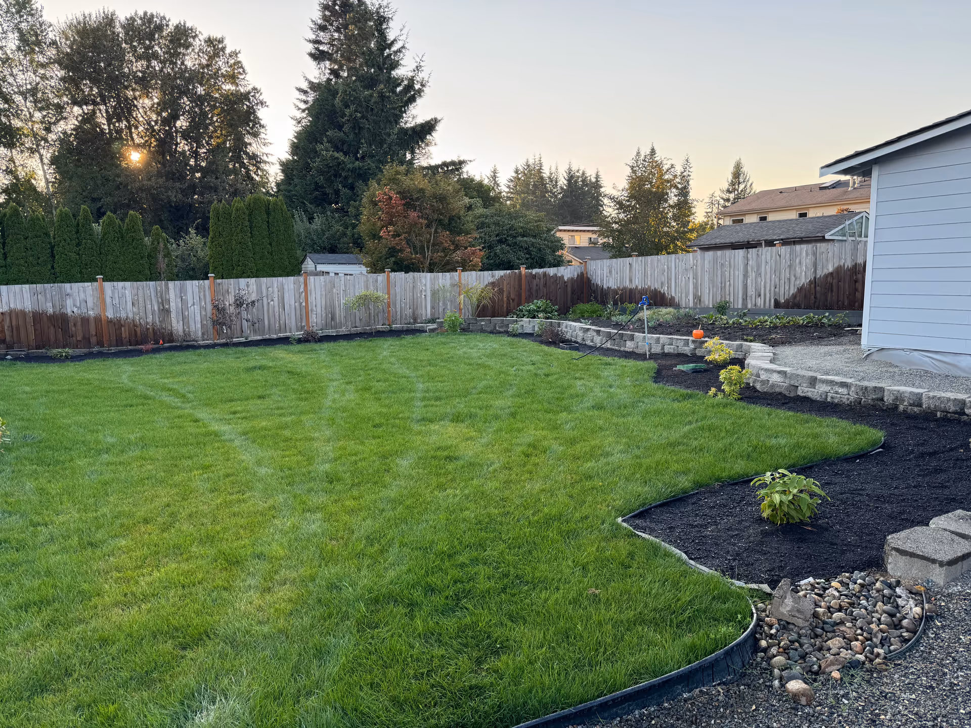 Exterior view of a fenced backyard with lush lawn and garden beds beside a house.
