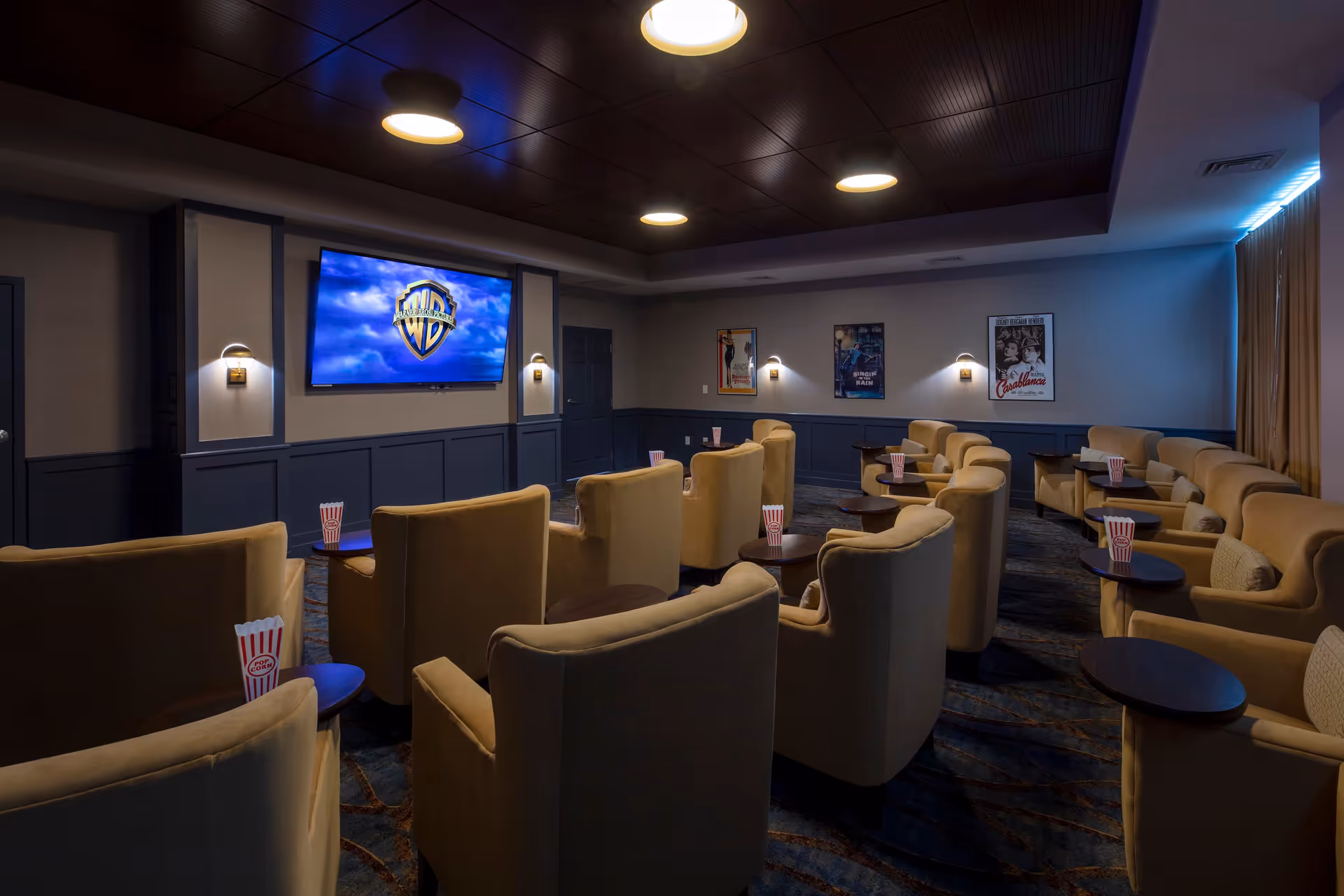 A cozy movie theater room with multiple rows of beige armchairs, each paired with a small round side table holding popcorn containers. A large flat-screen TV is mounted on the front wall displaying the Warner Bros. logo. The room has dim lighting with wall sconces and movie posters on the walls.
