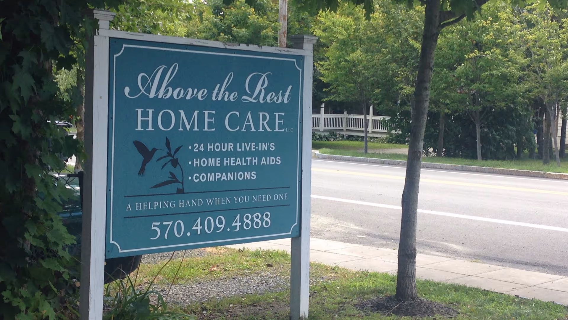A teal signboard for Above the Rest Home Care LLC beside a sidewalk and road.