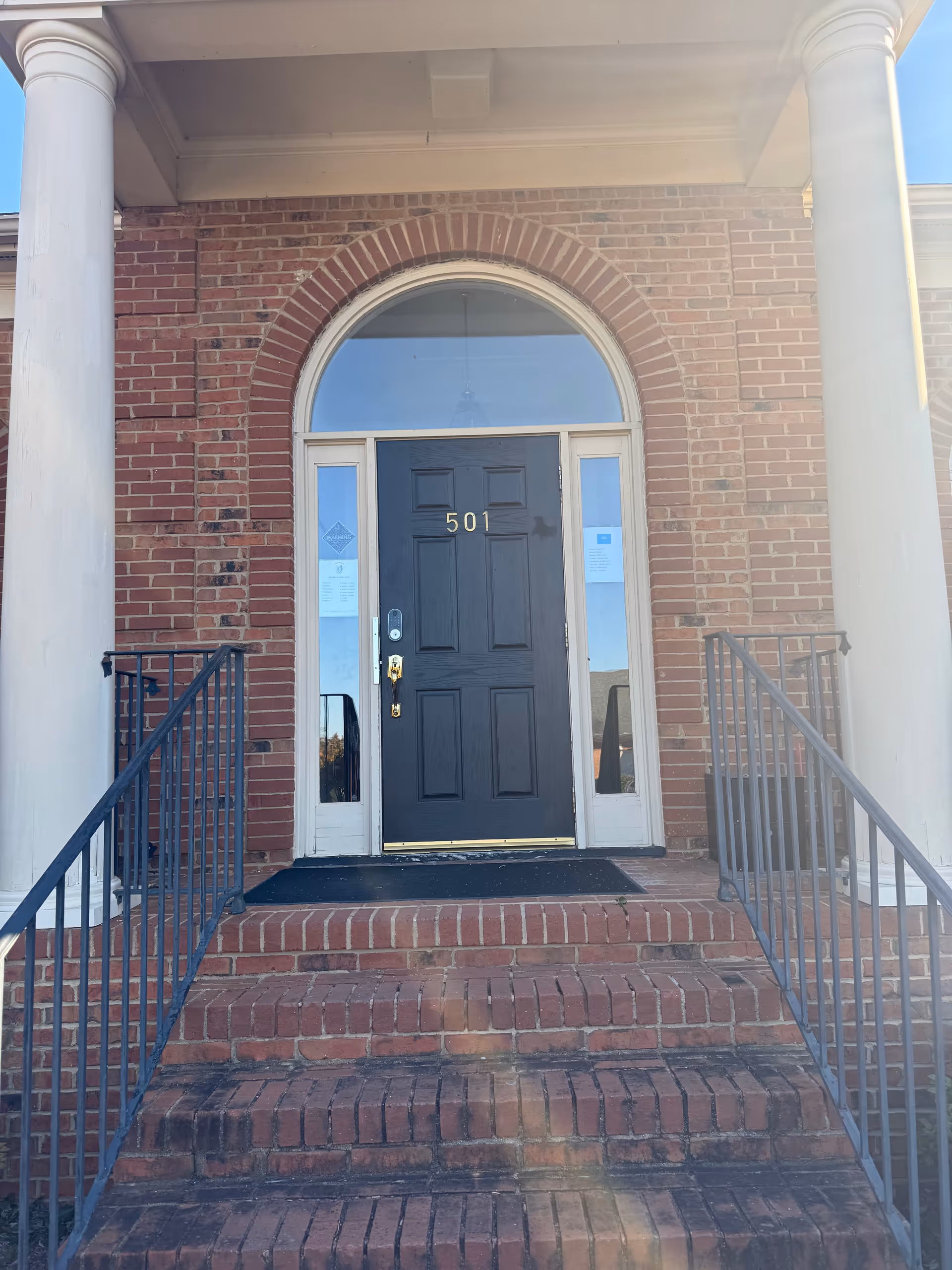 Brick building entrance with arched window, black door, steps, and white columns.