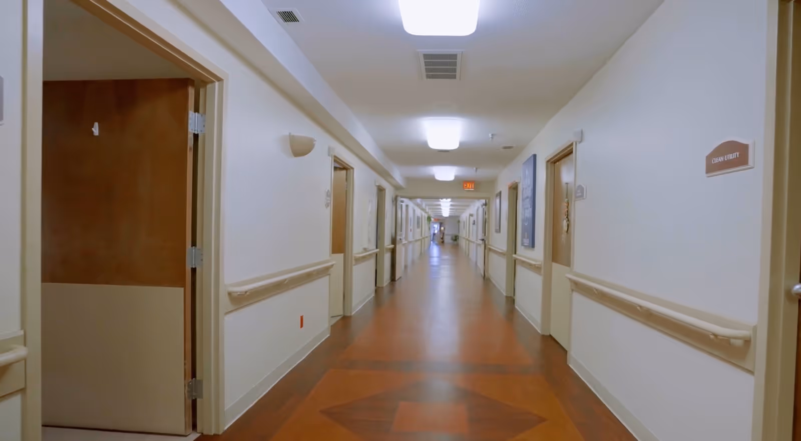 A long, well-lit hallway in a healthcare facility with wooden flooring and white walls. Doors line both sides of the corridor, some with signs such as 'Clean Utility'. Handrails run along the walls on both sides. The ceiling has rectangular light fixtures and ventilation grilles.