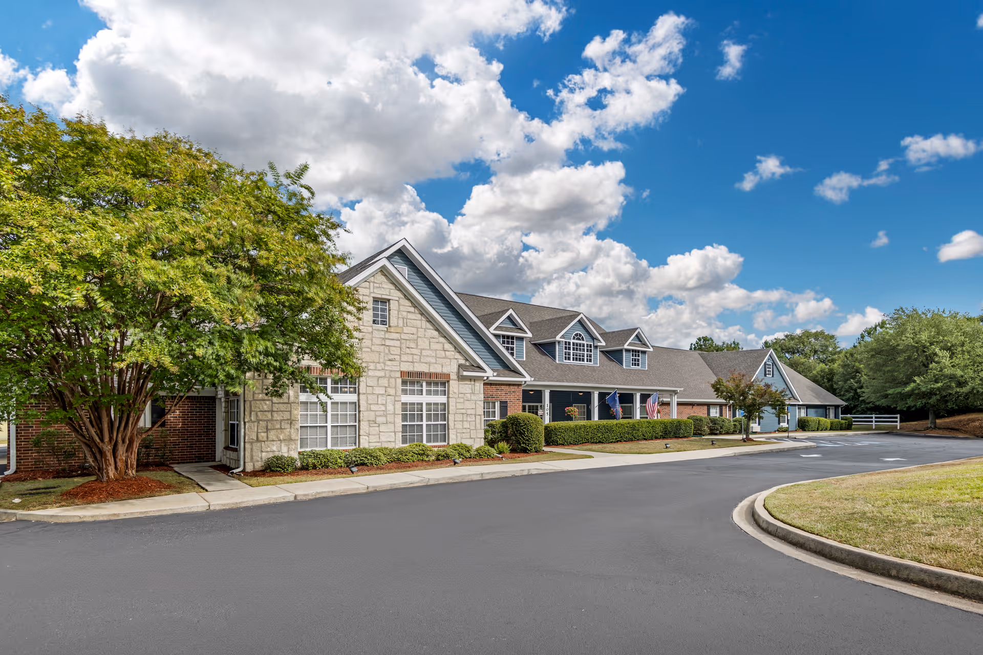 Front exterior of a single-story senior living building with stone and brick facade, landscaped shrubs, a curved driveway, and trees under a blue sky.