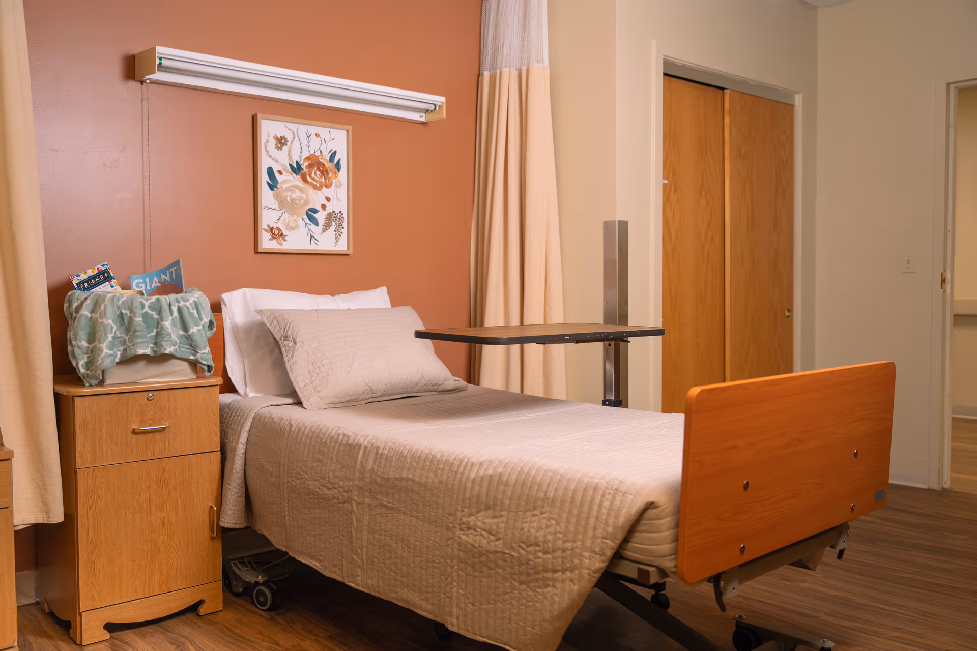 Hospital-style bed in a furnished patient room with a nightstand, curtains, and artwork.