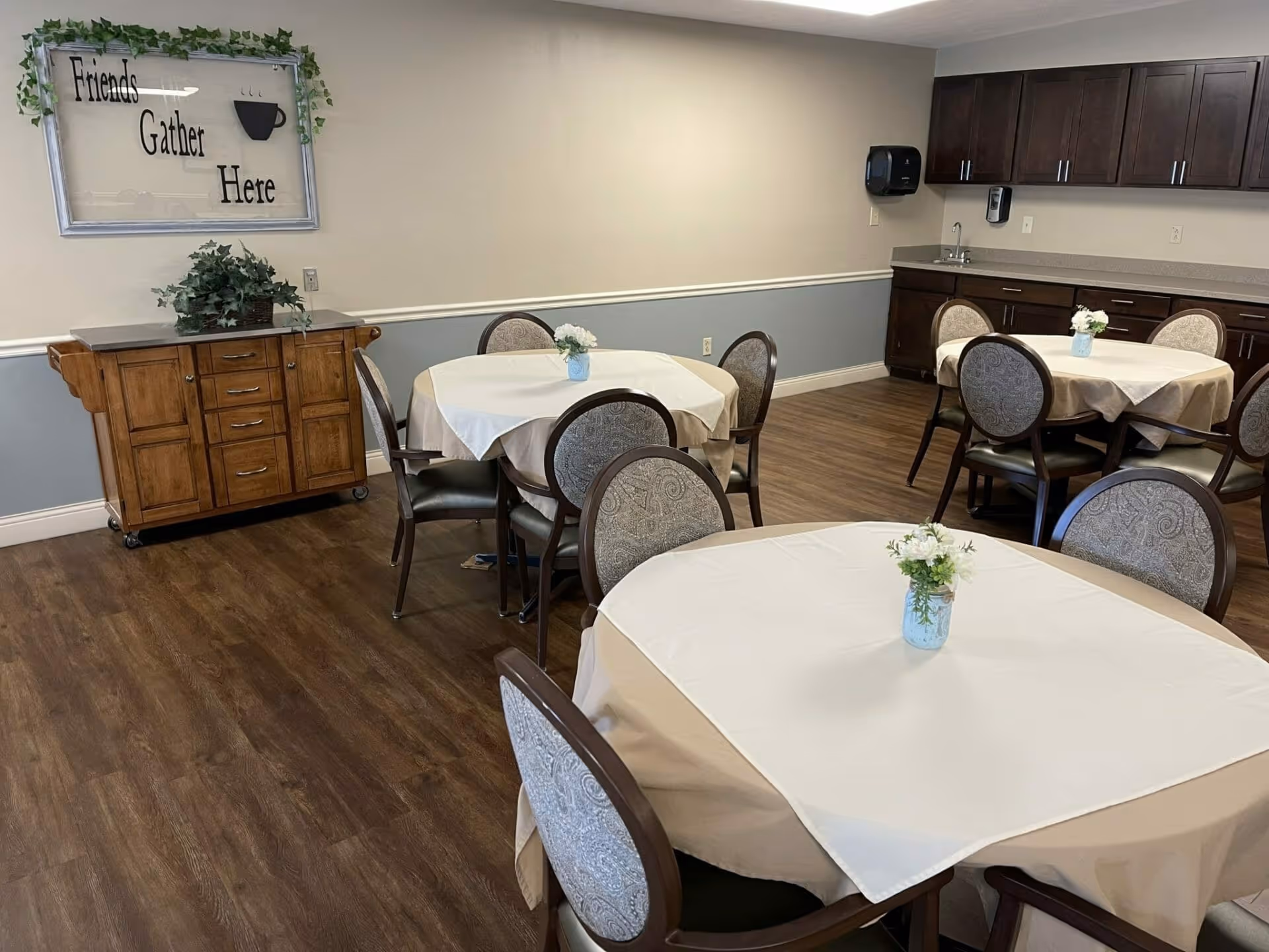Interior view of a dining room with round tables covered with beige tablecloths and white overlays, each table decorated with a small flower arrangement in a blue vase. The room has wooden flooring, light-colored walls with a blue-gray lower half, and dark wooden cabinets with a sink and paper towel dispenser. A wooden sideboard with a plant on top is against one wall, above which hangs a decorative sign that reads 'Friends Gather Here' with a coffee cup graphic.