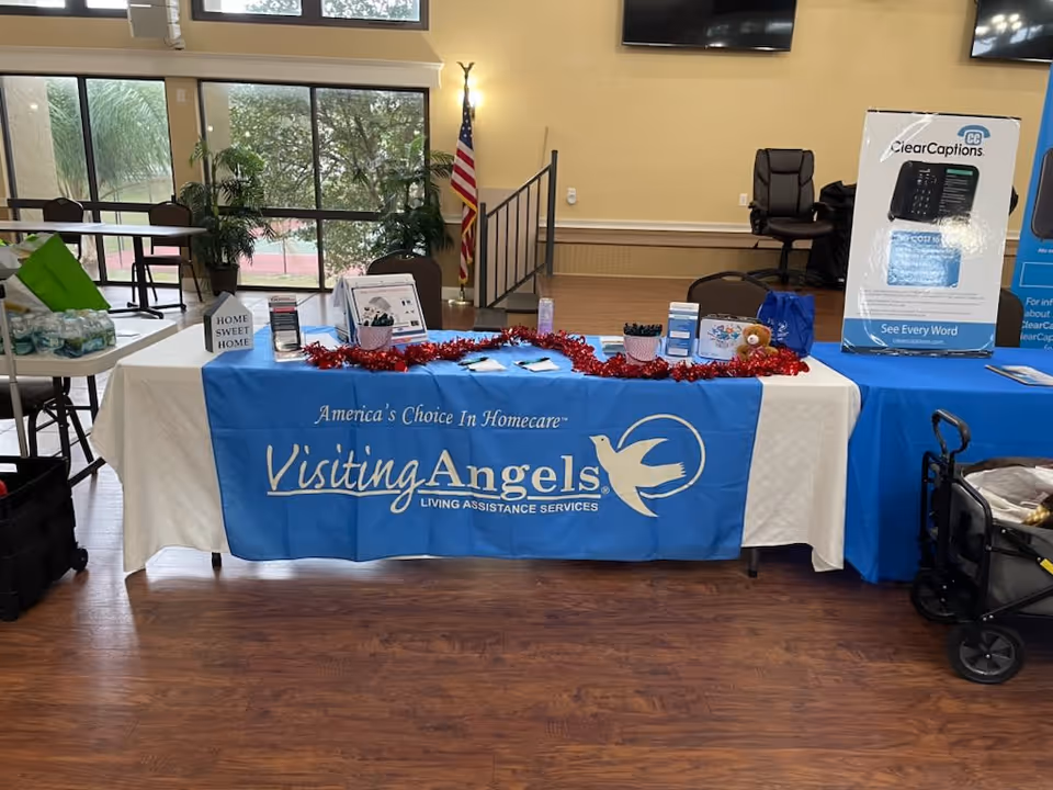 Indoor reception table with Visiting Angels banner, promotional materials, plants, and an American flag