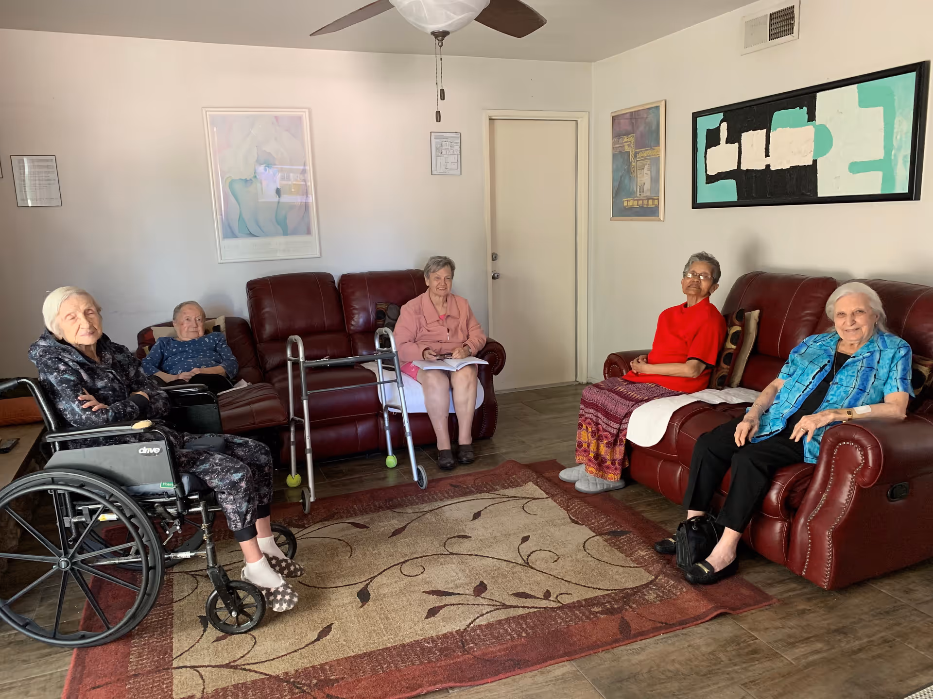 Five elderly women sitting in a living room with red leather sofas and a patterned rug. One woman is in a wheelchair, another has a walker in front of her, and the others are seated on sofas. The room has a ceiling fan, framed artwork on the walls, and a closed door in the background.