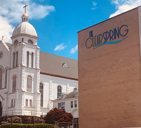 Brick facade labeled 'The Clearspring' beside a white church with a domed tower under a blue sky.