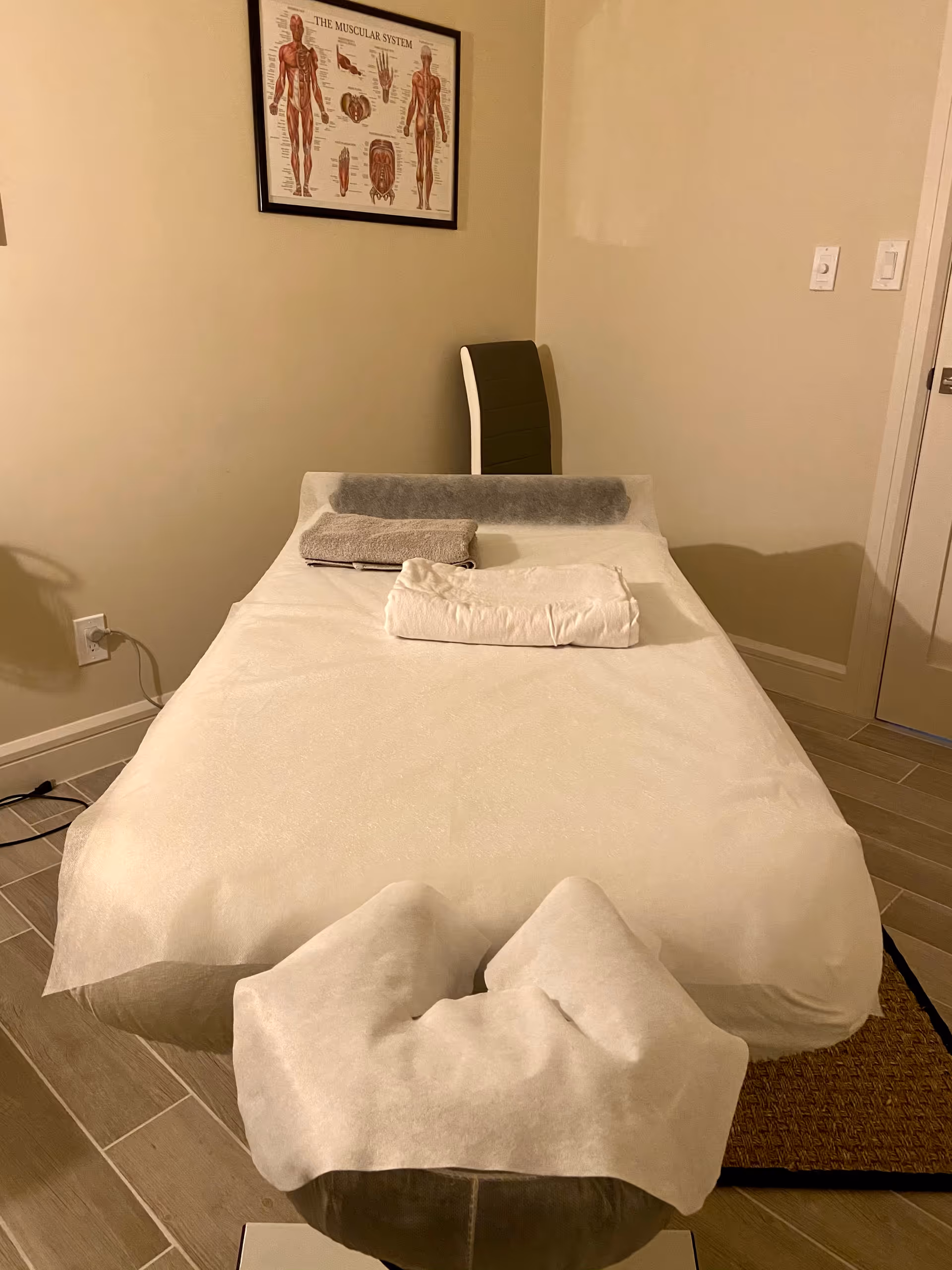 A massage table in a beige-walled treatment room with pillows and anatomy poster.