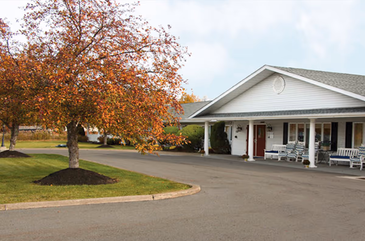 Driveway and entrance of a single-story senior living building with a covered porch, benches, and autumn trees.