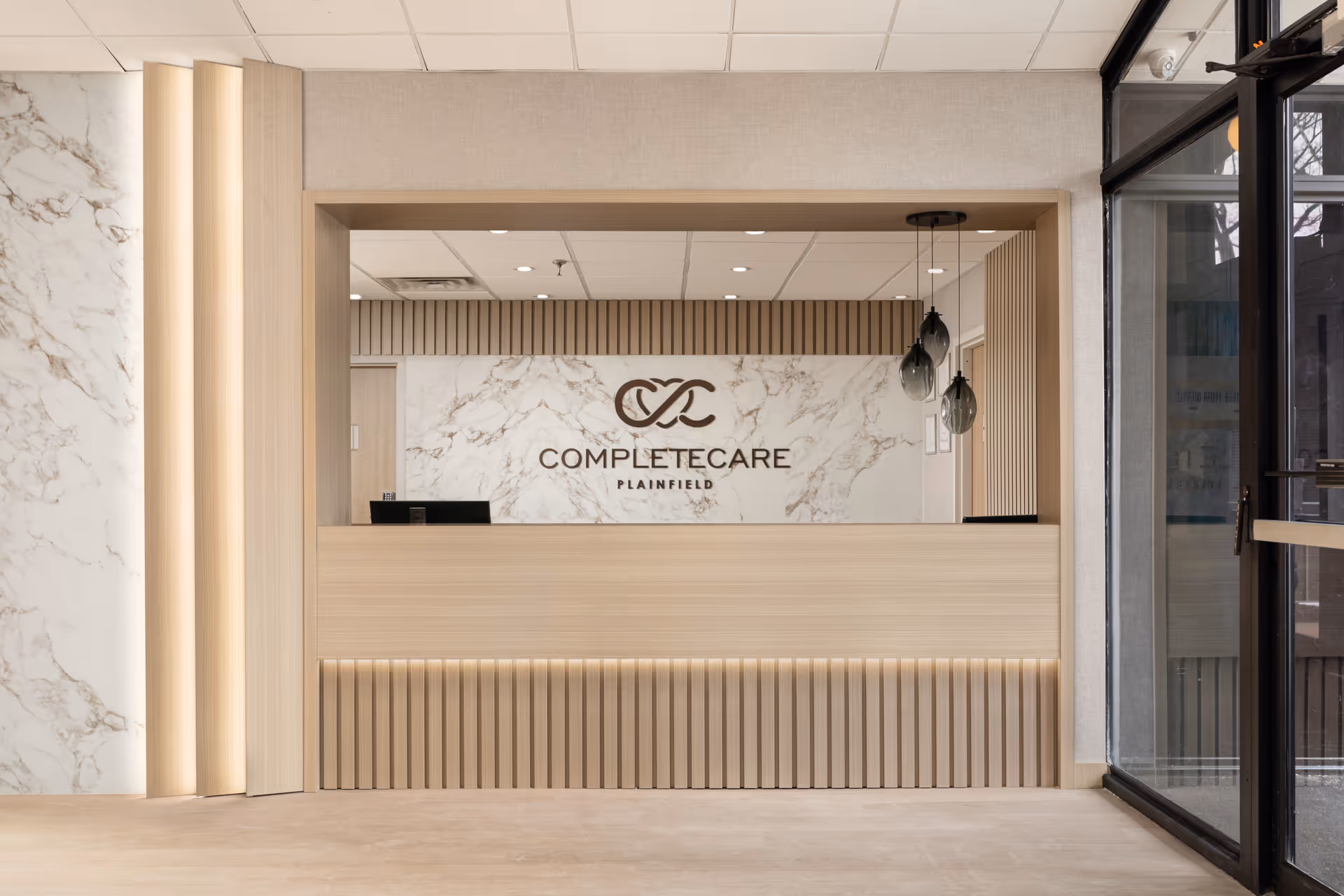 Modern lobby reception with marble wall, wooden counter, and pendant lights labeled CompleteCare Plainfield.