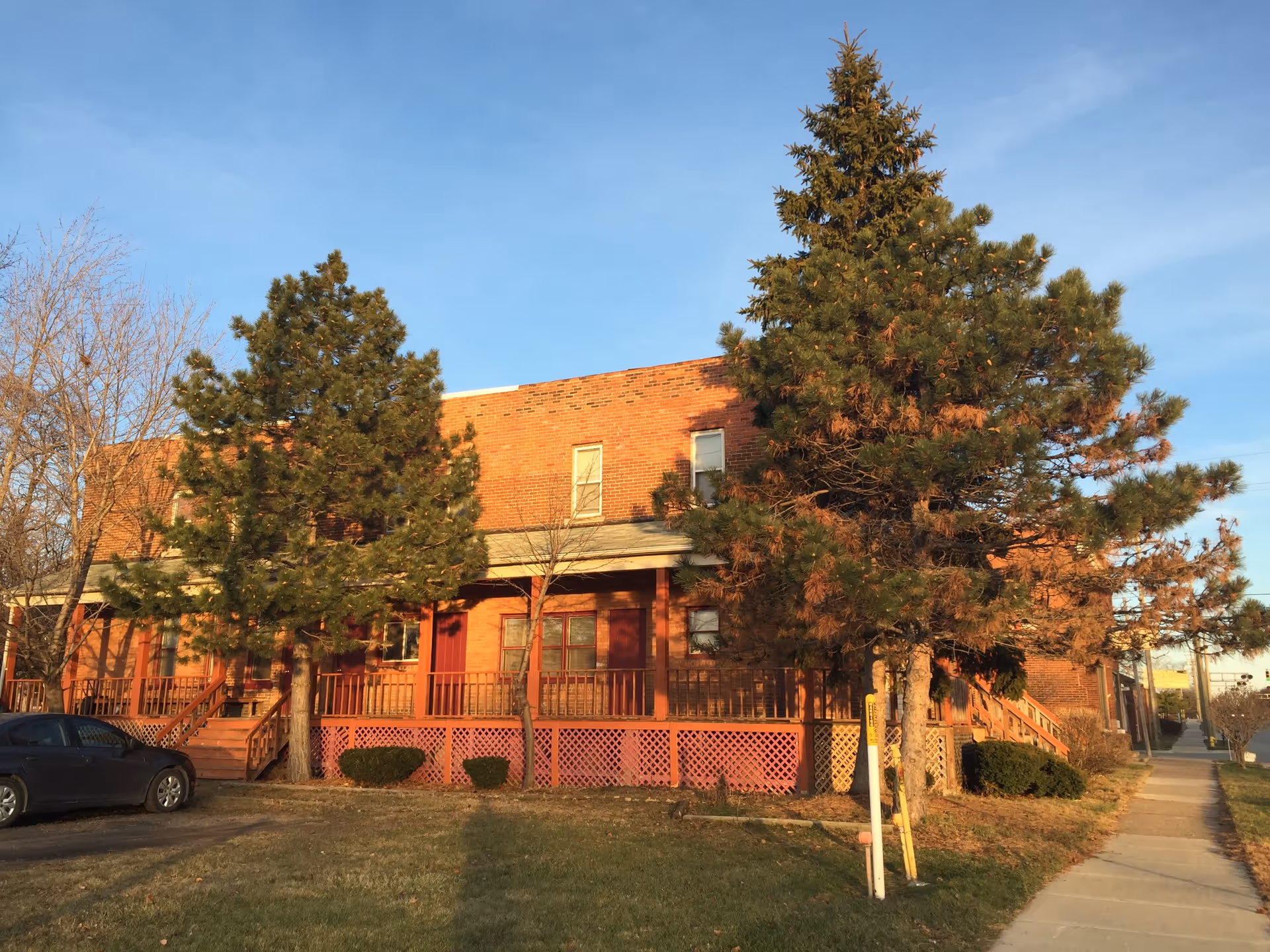 Brick senior living home with wraparound porch, large trees, and a sidewalk along the street.