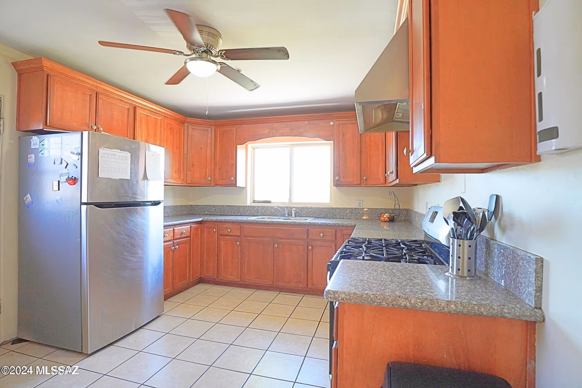 Bright kitchen with wooden cabinets, stainless refrigerator, stove, granite counters, and a ceiling fan.
