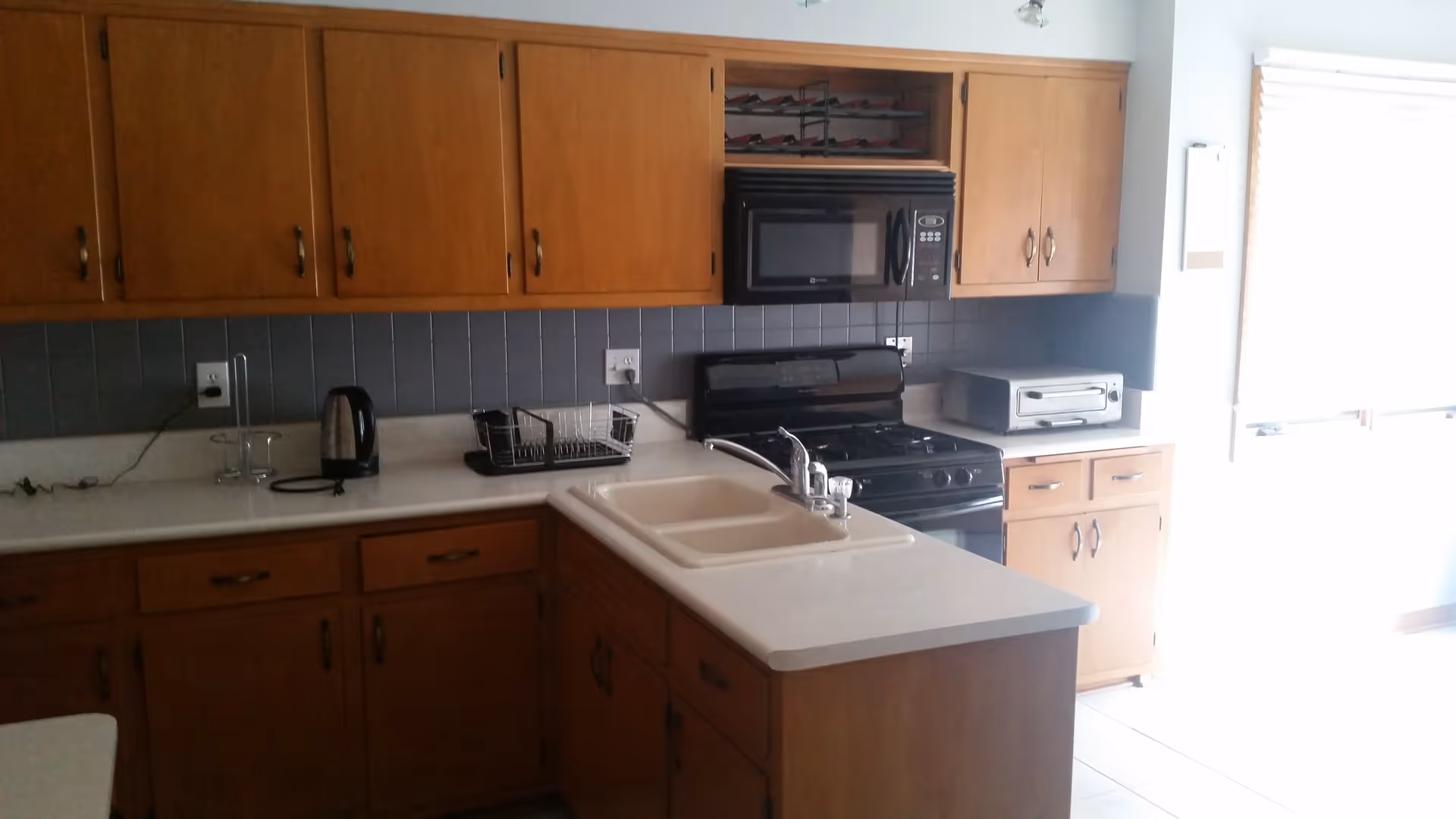 A kitchen with wooden cabinets, a double sink on a countertop island, a black microwave above a black stove, a toaster oven on the counter, a kettle, and a dish drying rack. There is a window with blinds letting in bright light on the right side.