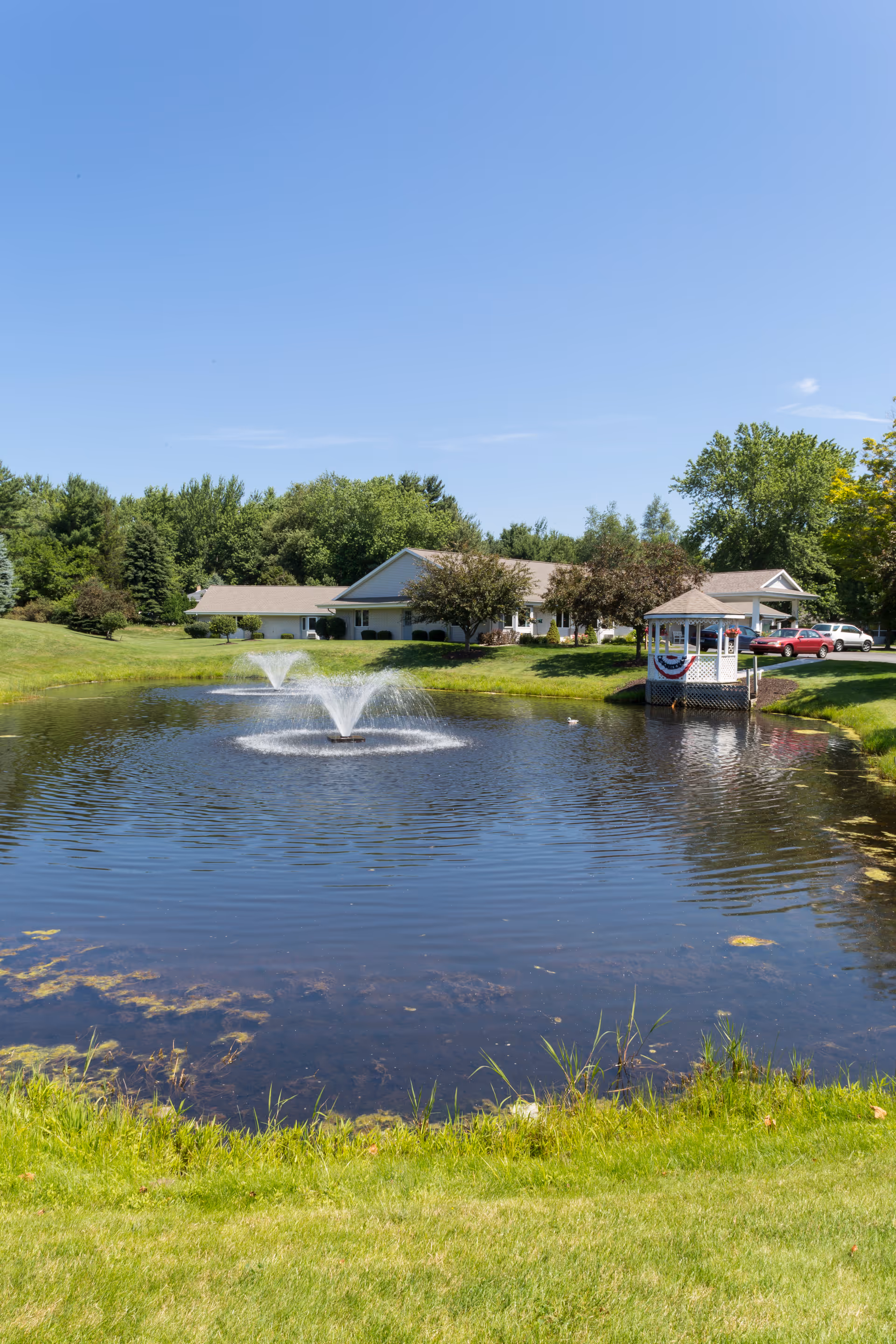 Outdoor view of a pond with fountains, a pavilion, and a low-slung senior living facility.