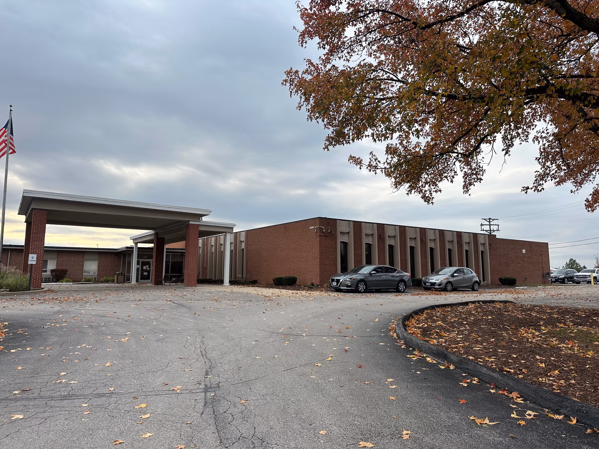 Single-story brick building with a covered entrance, parked cars, and an autumn tree in the foreground under a cloudy sky.