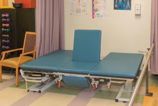 A physical therapy treatment room with a padded adjustable therapy table in teal color, a wooden chair with armrests, a rack holding colorful dumbbells, anatomical charts on the wall, and a hand sanitizer dispenser mounted on the wall.
