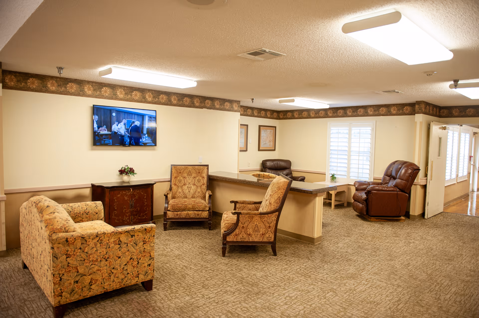 A cozy senior living facility common area with patterned upholstered chairs and a loveseat arranged around a small wooden cabinet. A flat-screen TV is mounted on the wall above the cabinet. The room has beige walls with a floral border near the ceiling, carpeted floor, and large windows with white shutters. There is a countertop partition separating the seating area from another section with additional chairs and a small table.