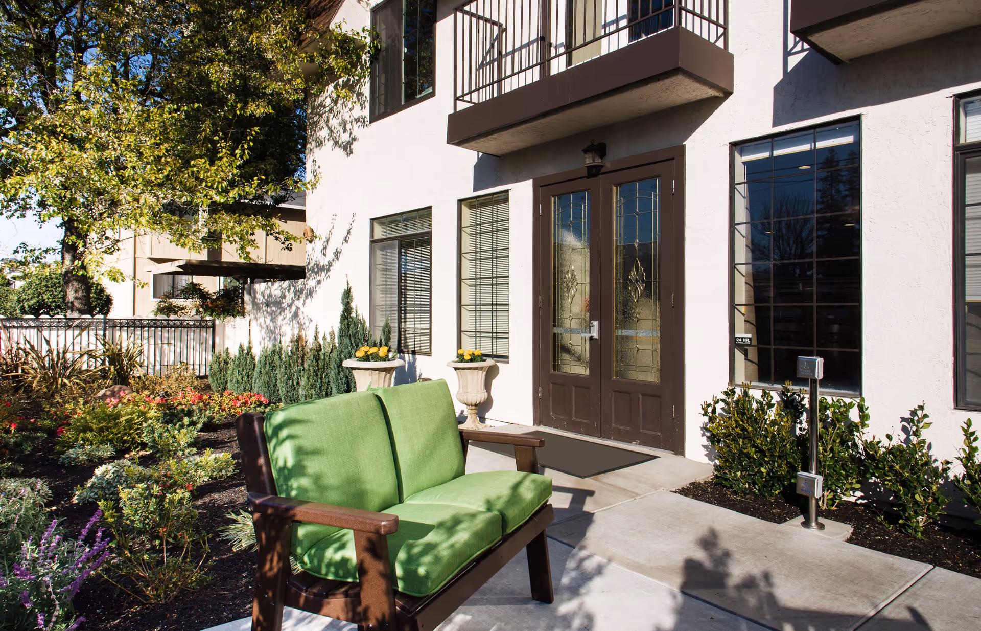 Outdoor patio area of Carefield Castro Valley Assisted Living and Memory Care with a green cushioned wooden bench, landscaped garden with various plants and flowers, and a building entrance featuring double glass doors and windows.