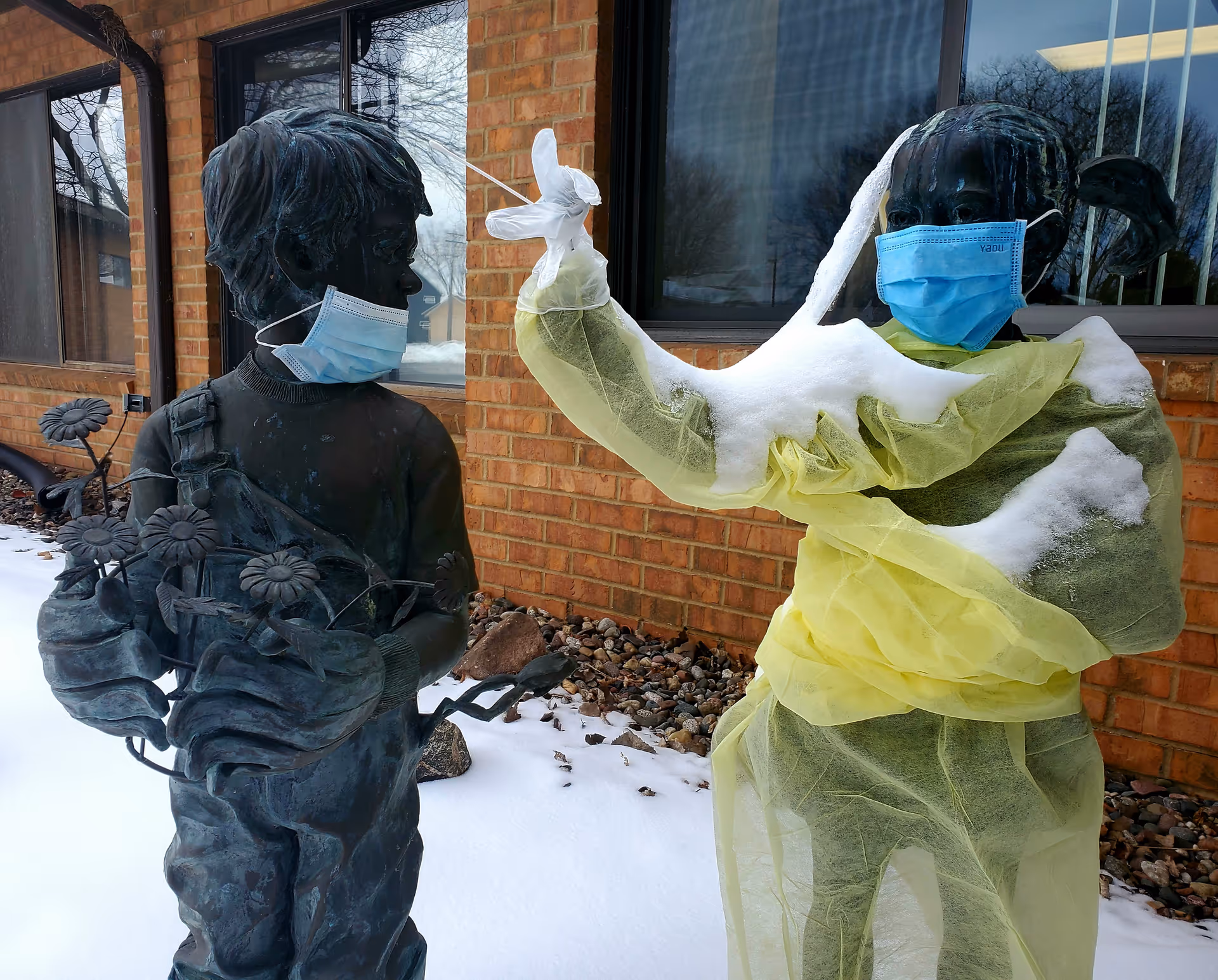 Two life-size statues wearing masks and yellow PPE outside a brick building.