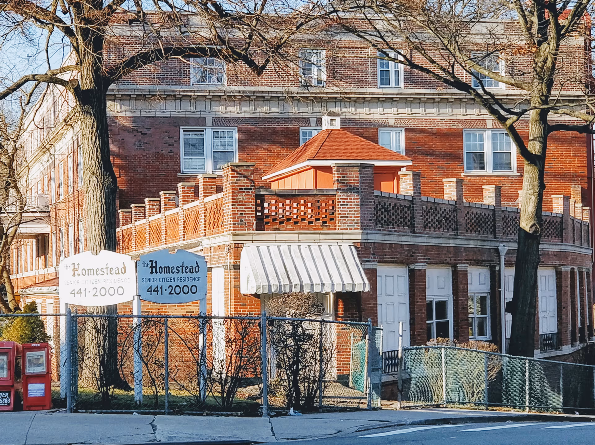 Brick senior residence building with front awning, two signs, and a fence.