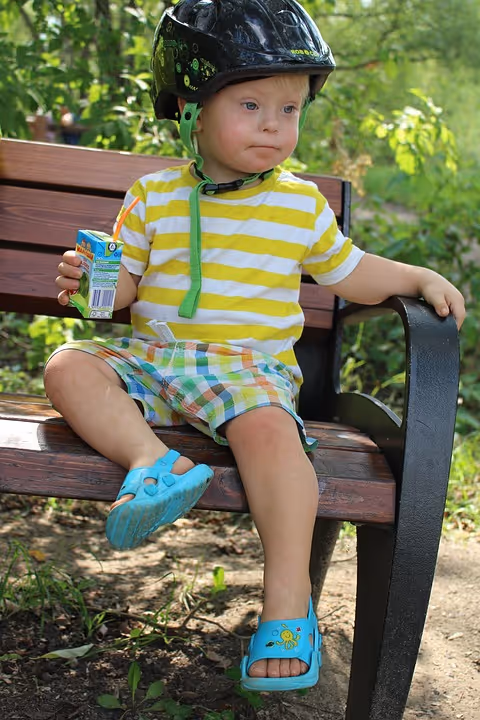 Young child wearing a helmet sits on an outdoor bench, holding juice box in a park.