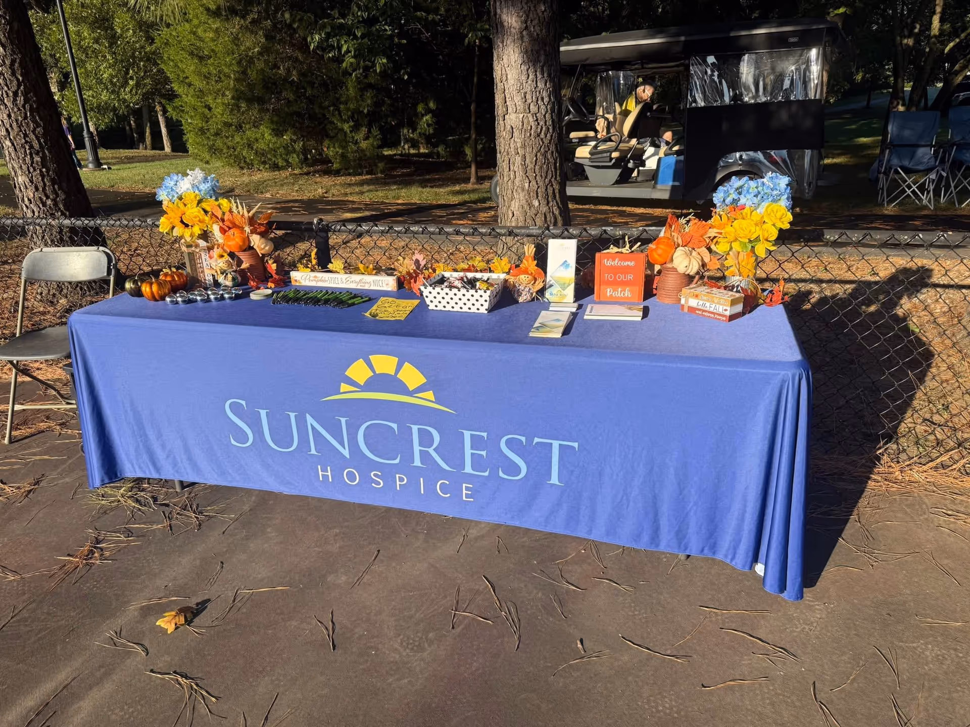 Outdoor event table with Suncrest Hospice banner, pumpkins, flowers, and chairs along fence.