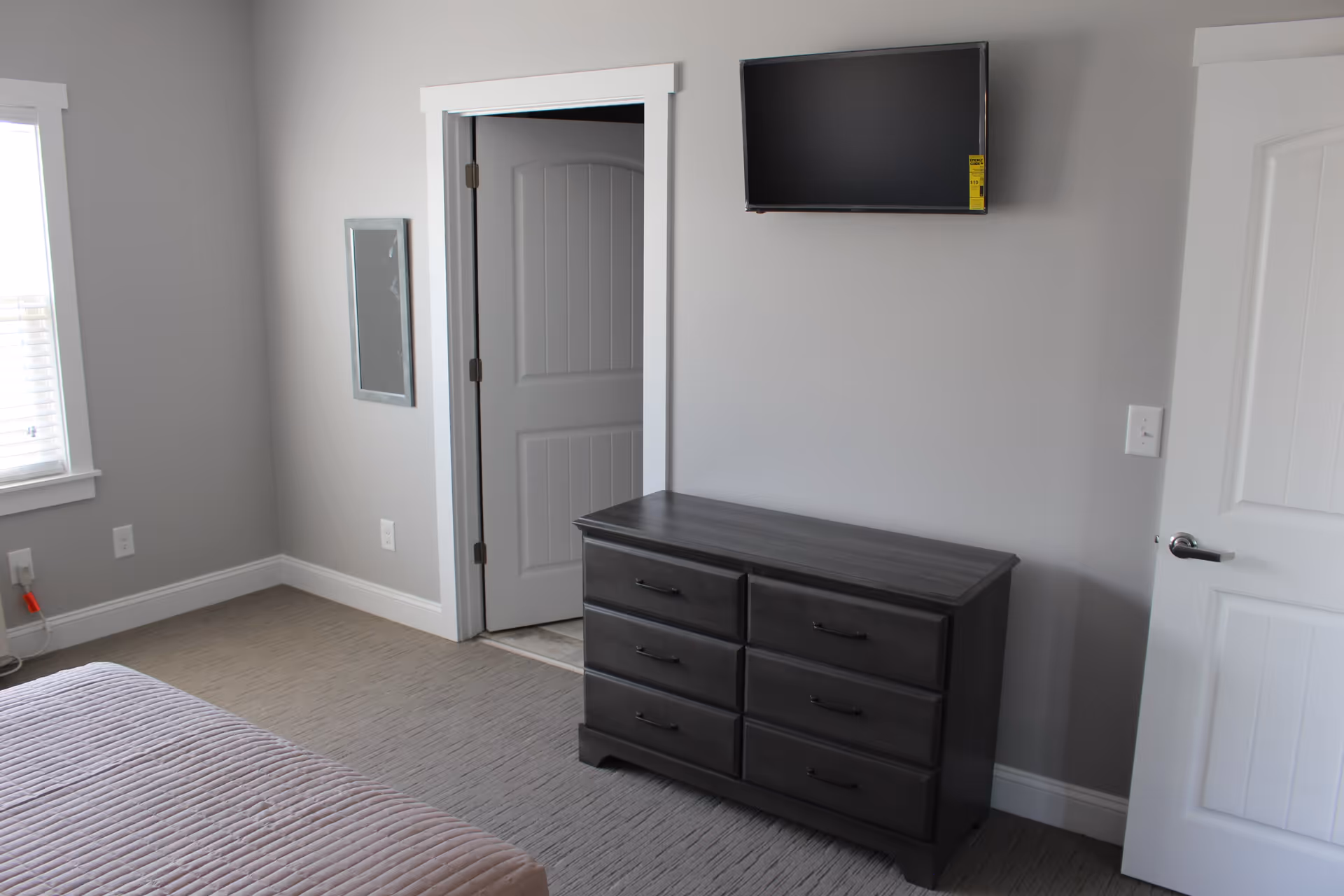 A bedroom with light gray walls and carpeted floor. There is a bed with a mauve quilt partially visible on the left side. A dark wooden dresser with six drawers is against the wall, and a flat-screen TV is mounted above it. Two white doors are visible, one slightly open leading to another room, and a window with blinds is on the left wall.
