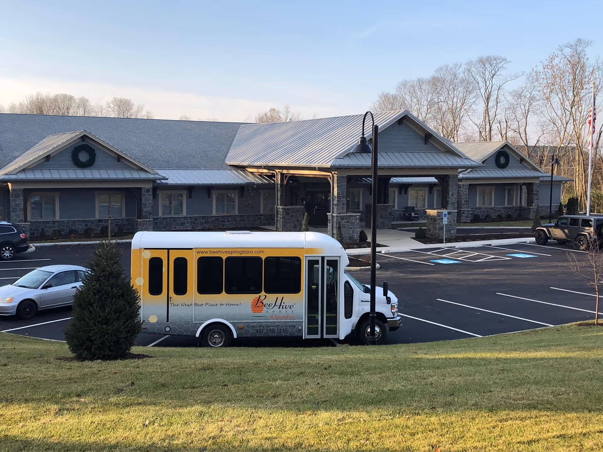 Front exterior of a senior living building with a yellow-and-white shuttle bus parked in the lot in front.