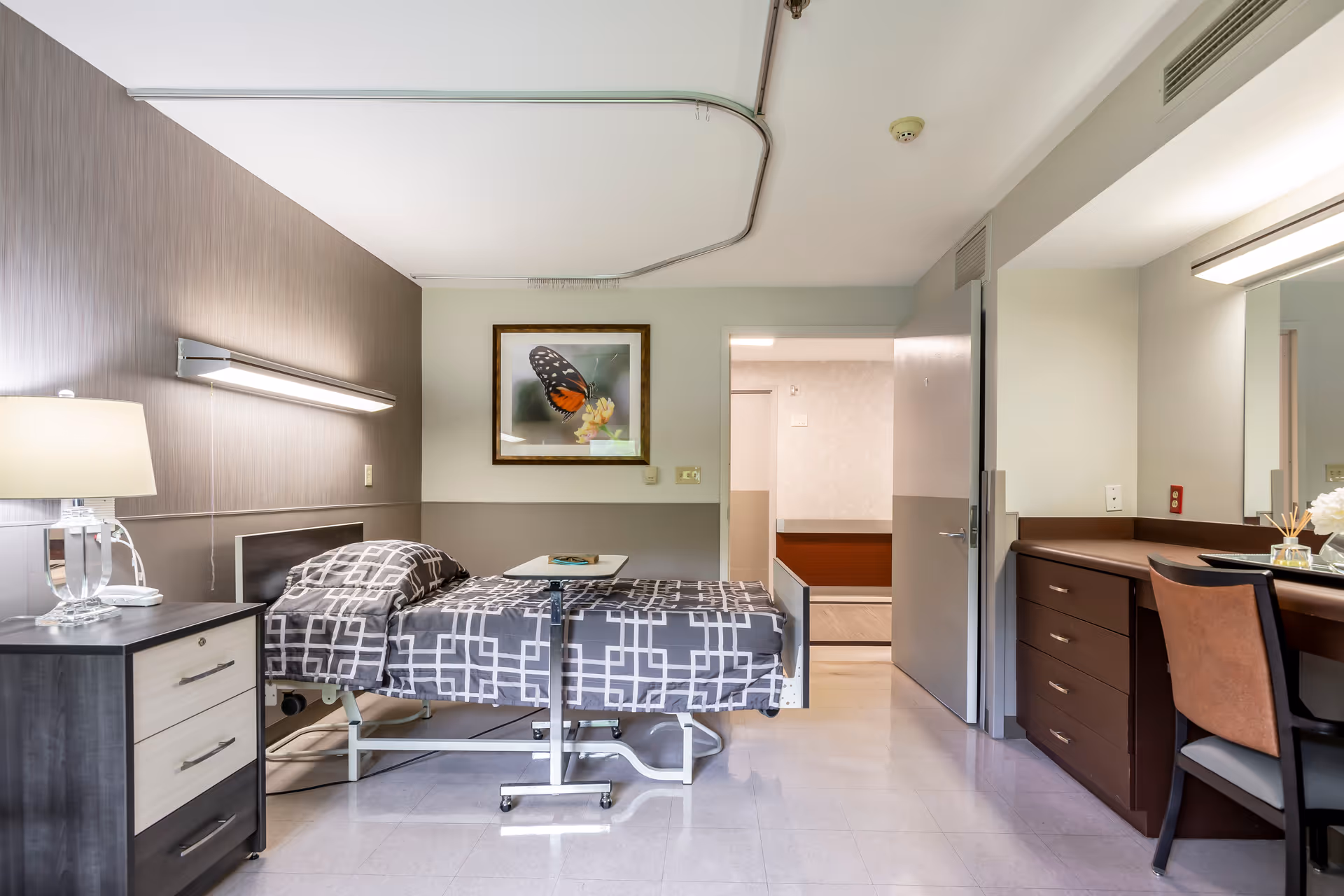 Hospital-style bedroom in a senior living facility with a bed, desk, and dresser.
