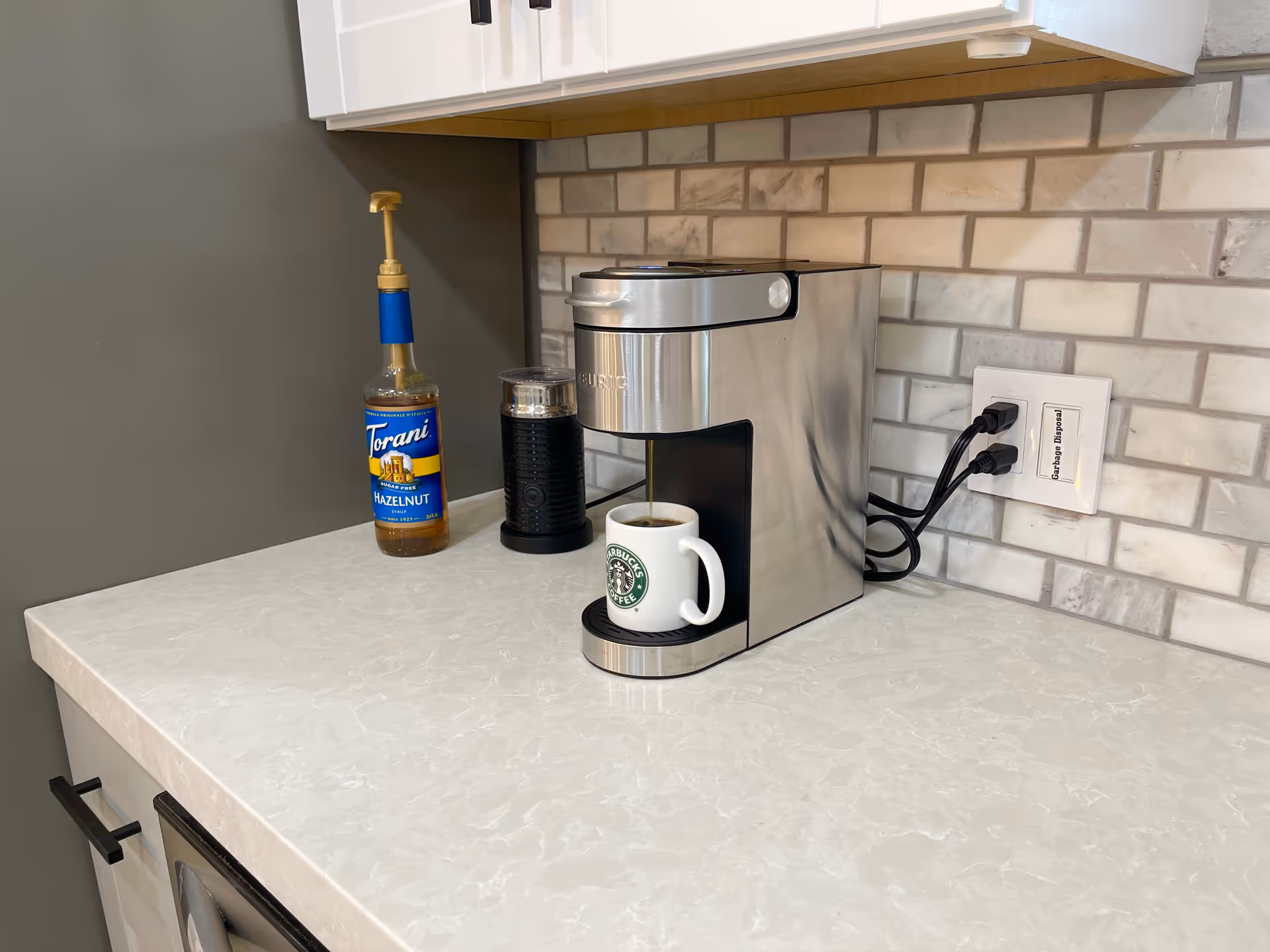 Stainless coffee machine on a marble counter with a mug and syrup bottle in kitchen.