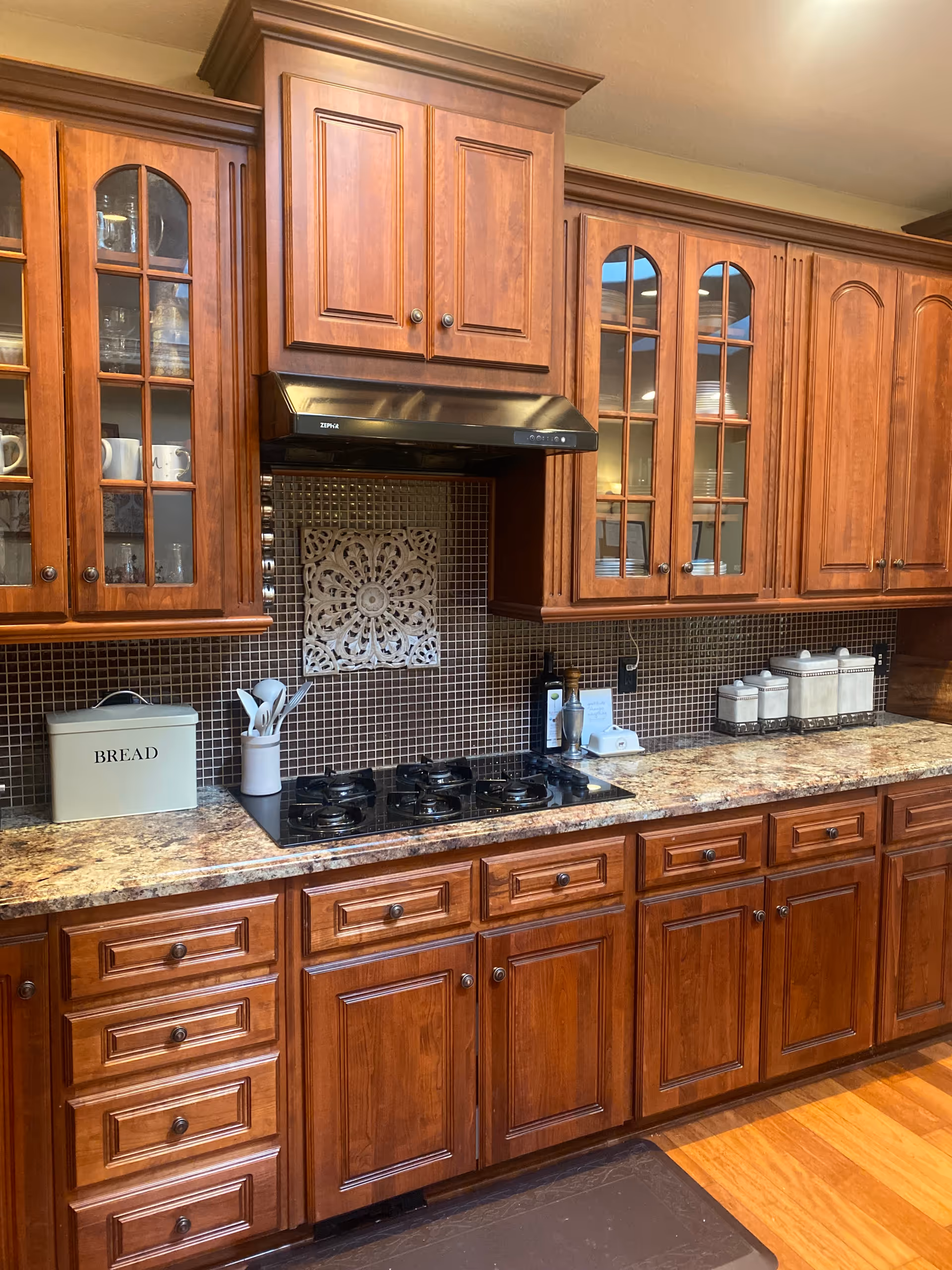 A kitchen with wooden cabinets and drawers, a granite countertop, a built-in gas stove with four burners, a stainless steel range hood, and a tiled backsplash with a decorative tile centerpiece. On the countertop, there is a bread box, a utensil holder with white utensils, and three white canisters with lids.