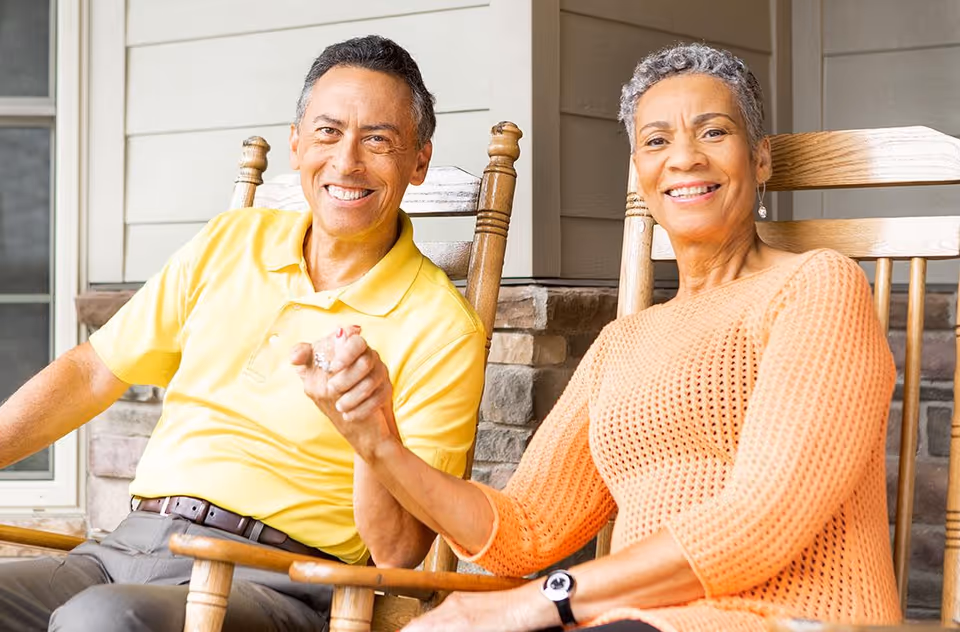 An elderly couple sitting on wooden rocking chairs on a porch, holding hands and smiling at the camera. The man is wearing a yellow polo shirt and gray pants, and the woman is wearing an orange knit sweater and a watch. The background shows a stone wall and siding of the building.