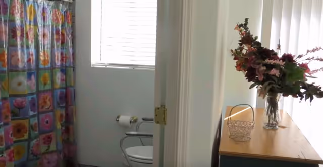 A bathroom with a toilet, grab bars, bright shower curtain, and a dresser topped with flowers.