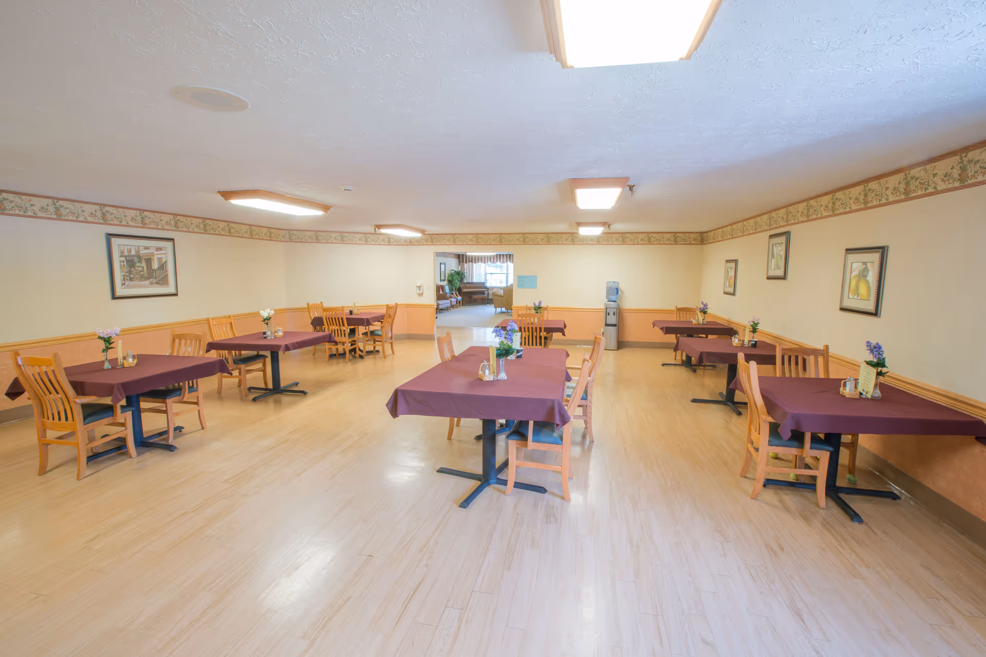A dining room with several wooden tables covered with purple tablecloths, each table surrounded by wooden chairs. Small flower arrangements and condiments are placed on the tables. The room has light-colored walls with a floral border near the ceiling, framed pictures on the walls, and a water dispenser in the back corner. The floor is light wood, and the ceiling has rectangular light fixtures.