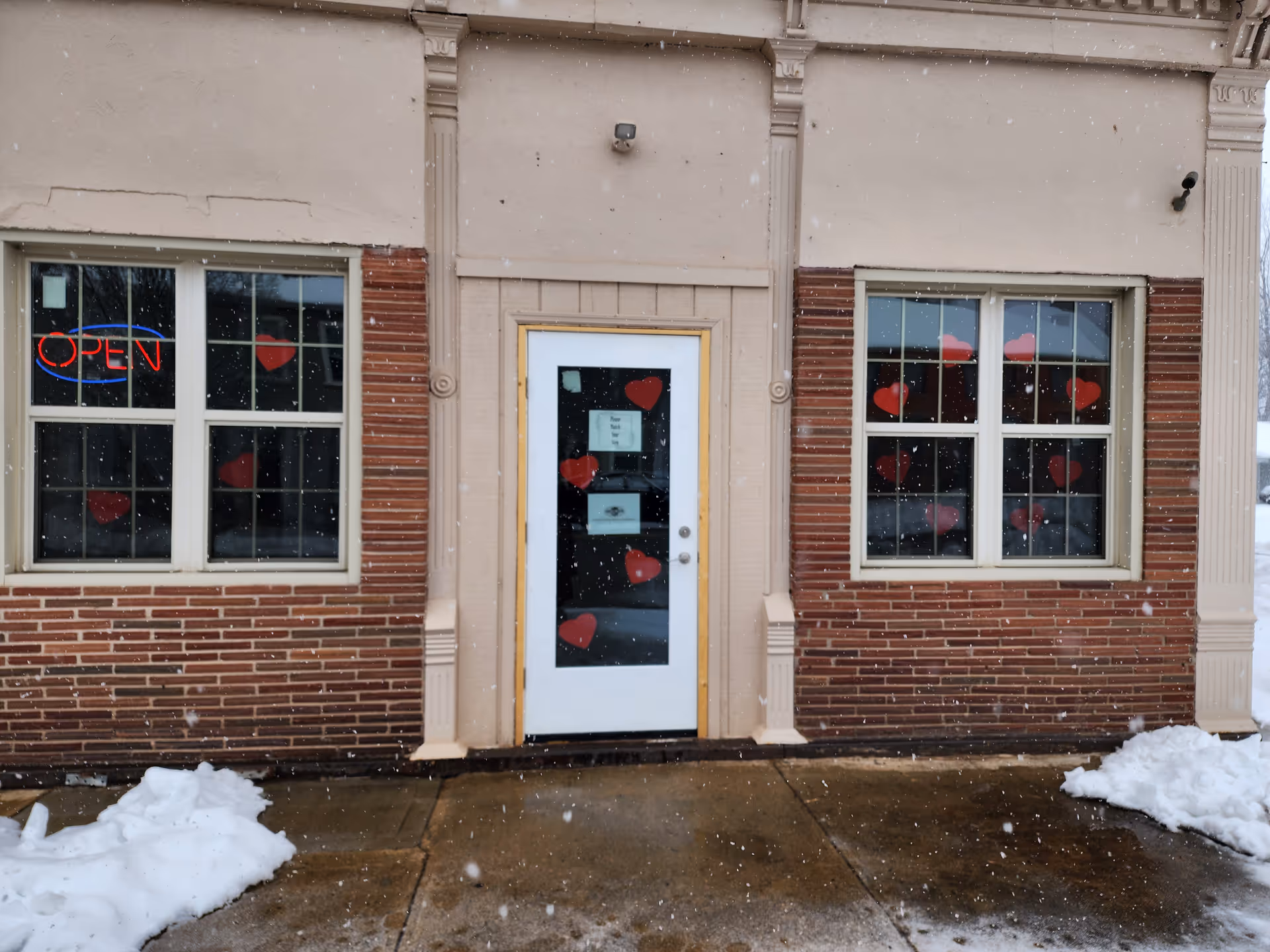 Front view of a brick building with doors and windows decorated with red hearts in snowfall.