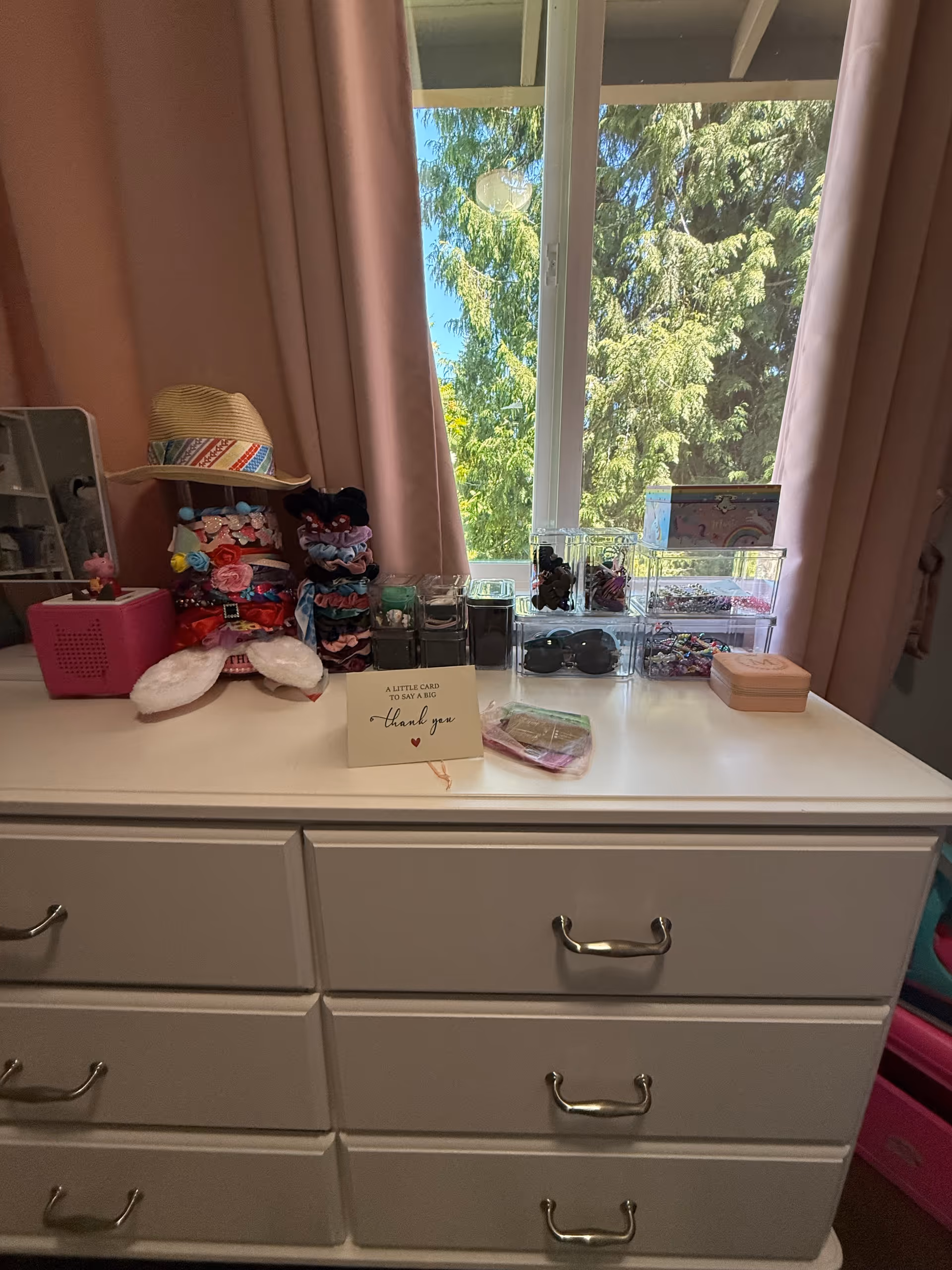 White dresser with drawers, cosmetics and accessories, beside a window with pink curtains.