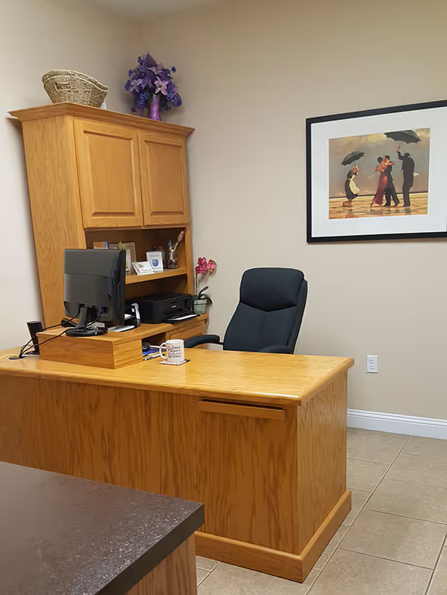 Beige-walled office with wooden reception desk, computer, cabinet, and framed artwork.