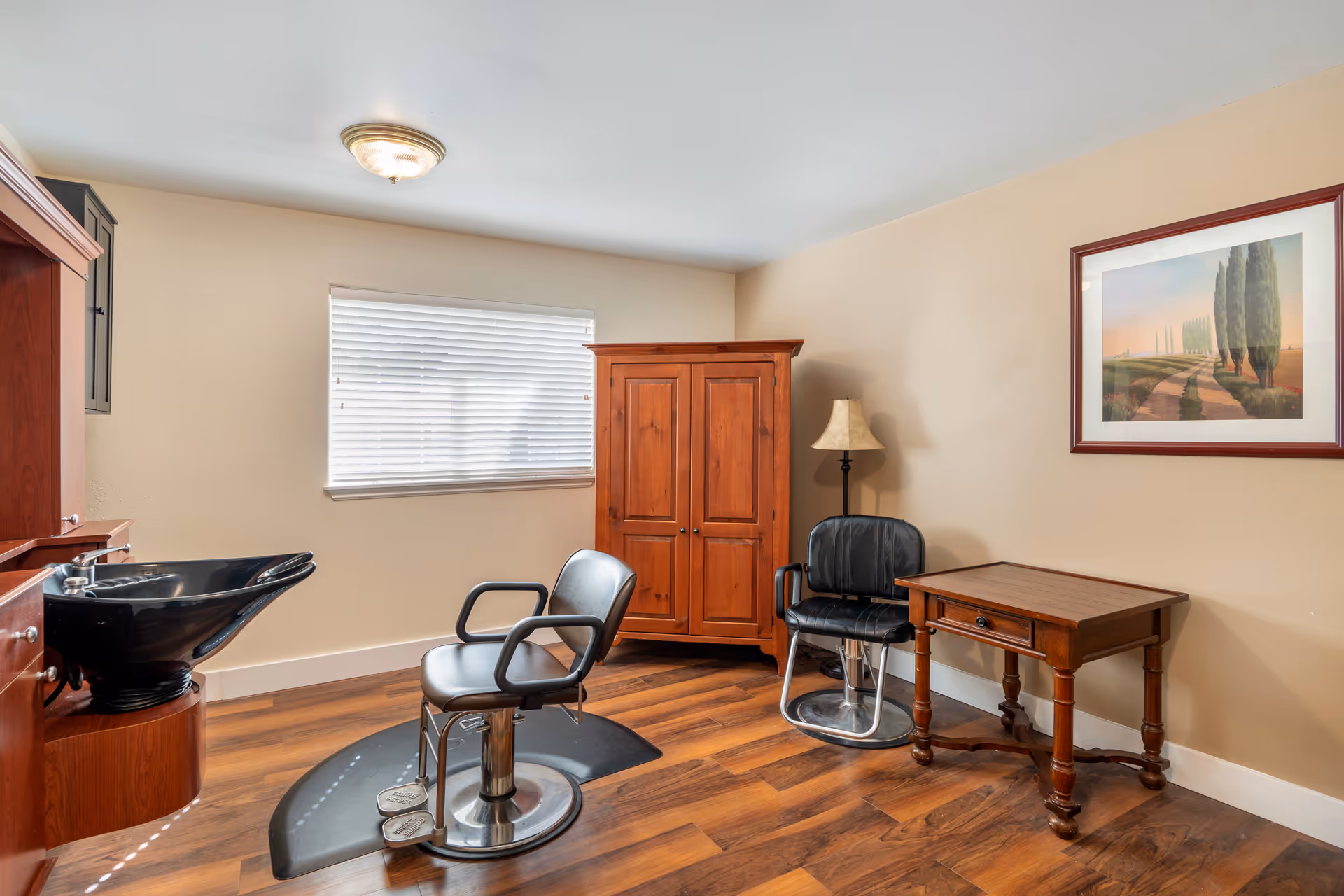A small salon room with two black salon chairs, a wooden cabinet, a wooden table, a floor lamp, and a framed landscape painting on the beige wall. There is a window with white blinds and a black salon sink on the left side.