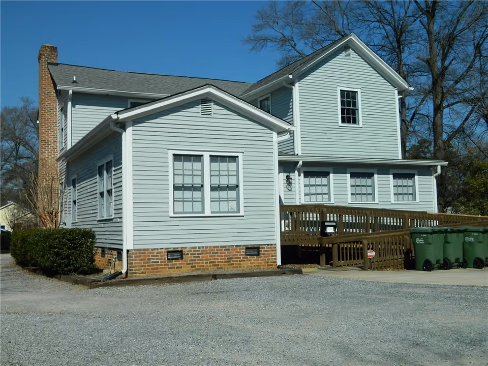 Two-story light-gray building with brick base, multiple windows, and a wooden ramp exterior.